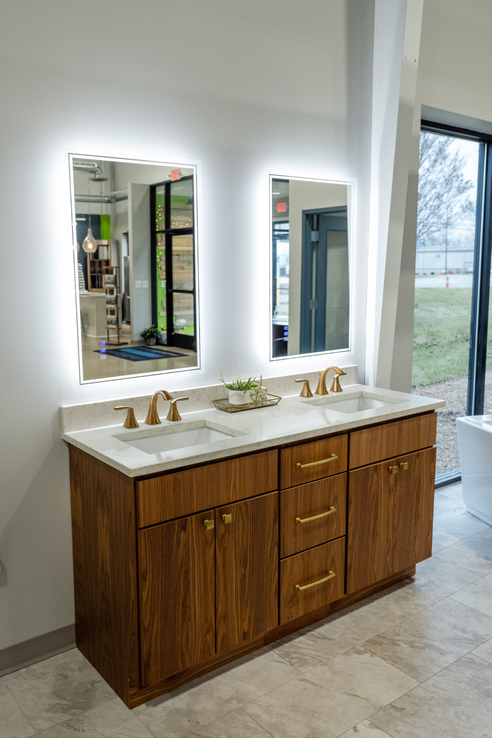 A modern bathroom vanity with a wood finish, dual sinks, gold faucets and handles, a small plant in the center, and two illuminated rectangular mirrors above—perfect for elevating your next bath remodel—set near large windows.