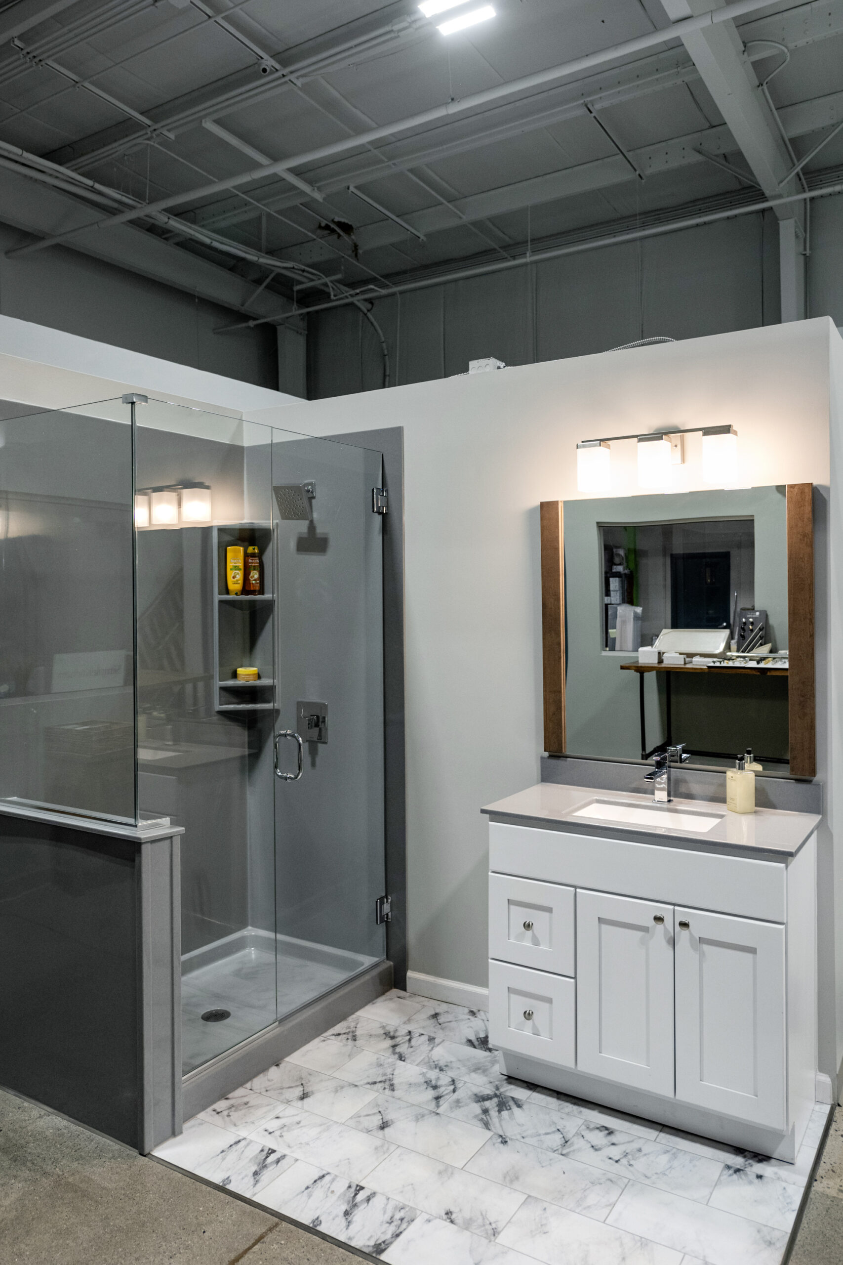 A modern bath remodel display featuring a glass-enclosed shower with built-in shelves, a white vanity with sink and mirror, and marble-patterned floor tiles under bright overhead lighting.