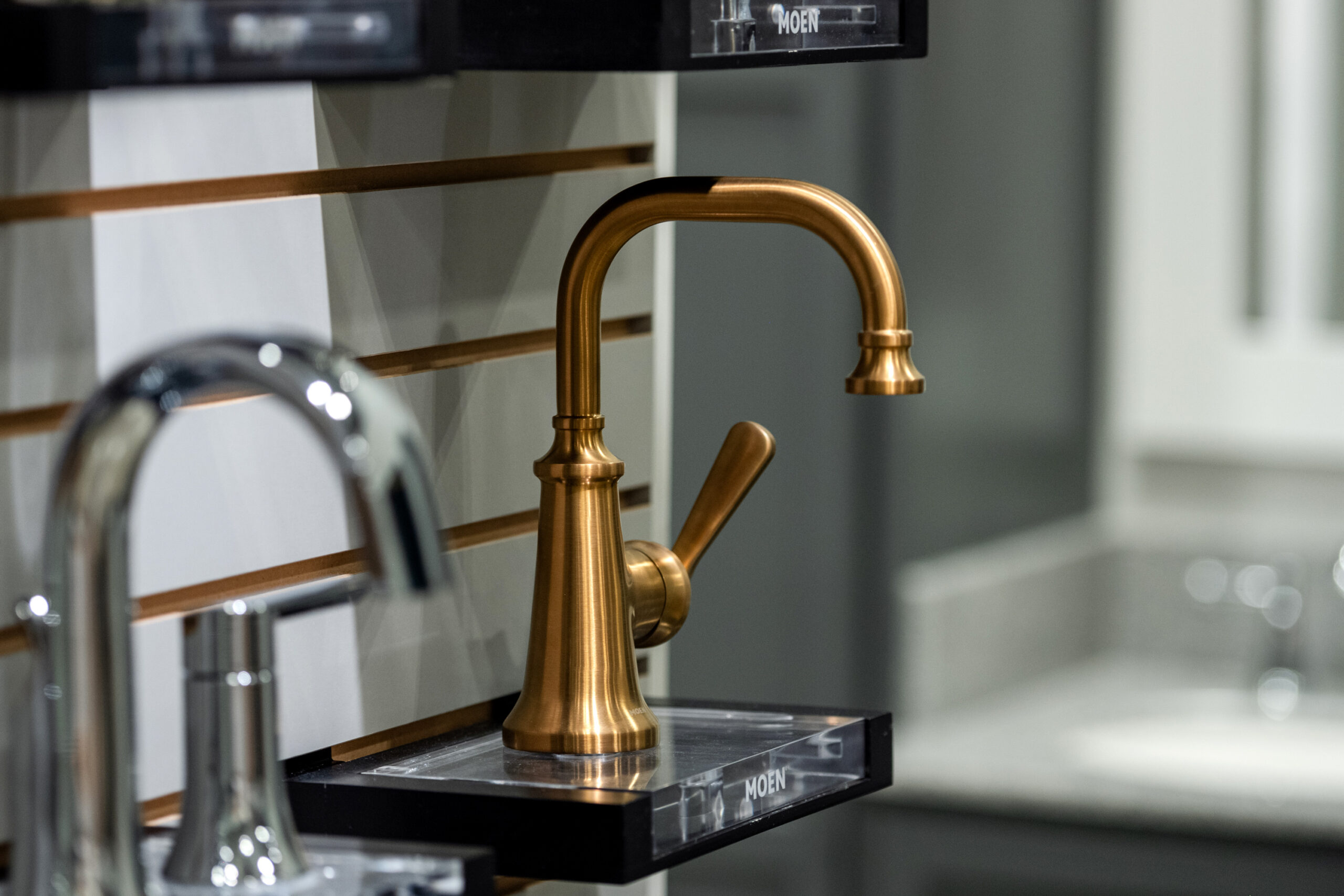A modern gold faucet with a curved spout and single lever handle is displayed on a showroom wall with horizontal slats—an inspiring choice for your next kitchen remodel. Another silver faucet is partially visible to the left.