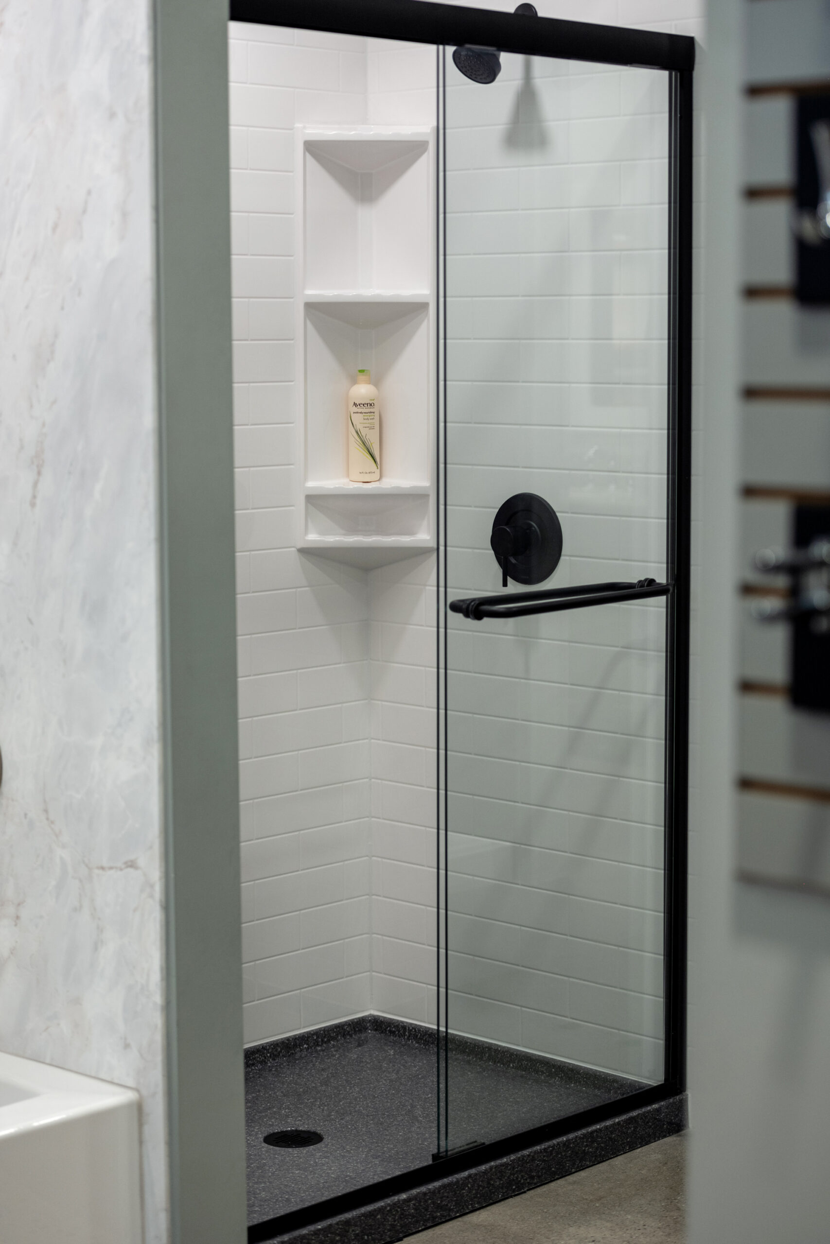 A modern shower, perfect for any bath remodel or bathroom renovation, with white subway tile walls, a glass door, black hardware, two built-in corner shelves, and a bottle of shampoo on the top shelf.