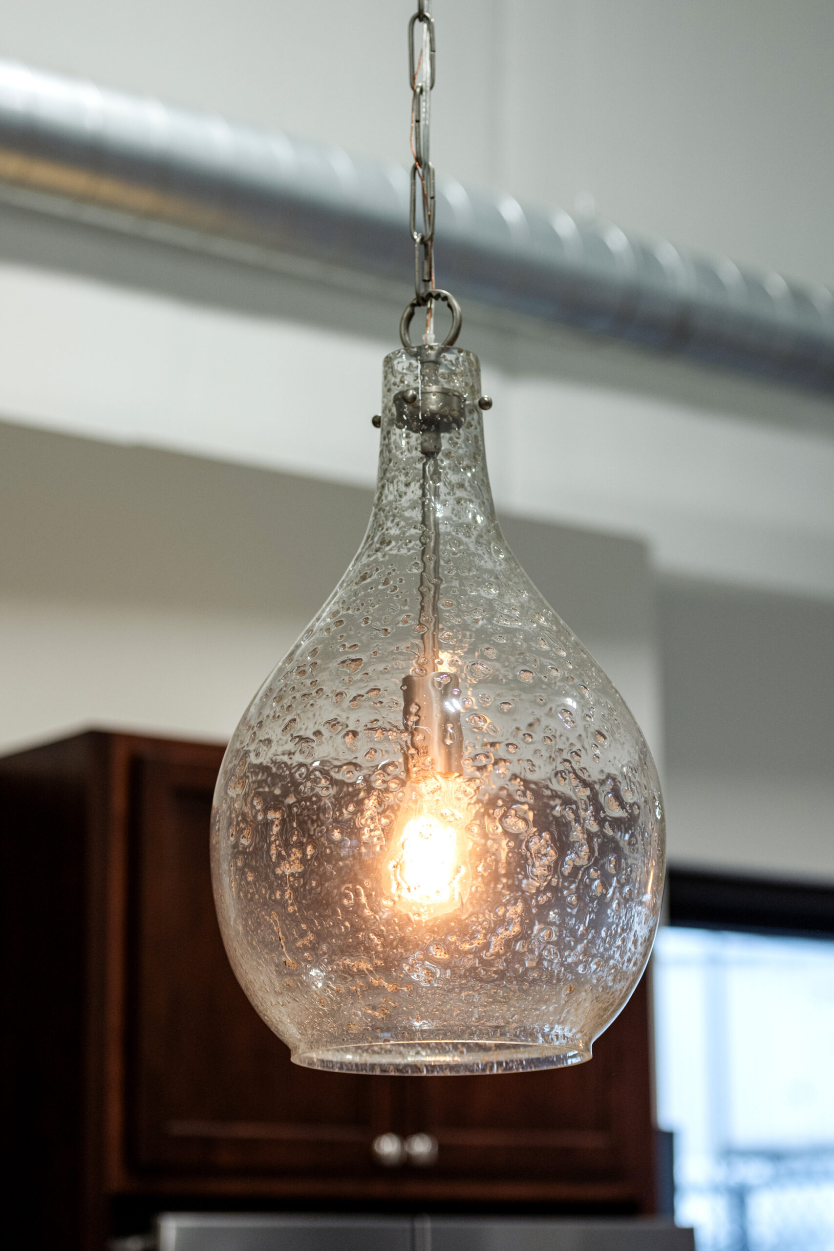 A teardrop-shaped glass pendant light with a textured, bubbled surface hangs from the ceiling, illuminating a modern kitchen renovation with visible cabinets and a metal air duct in the background.