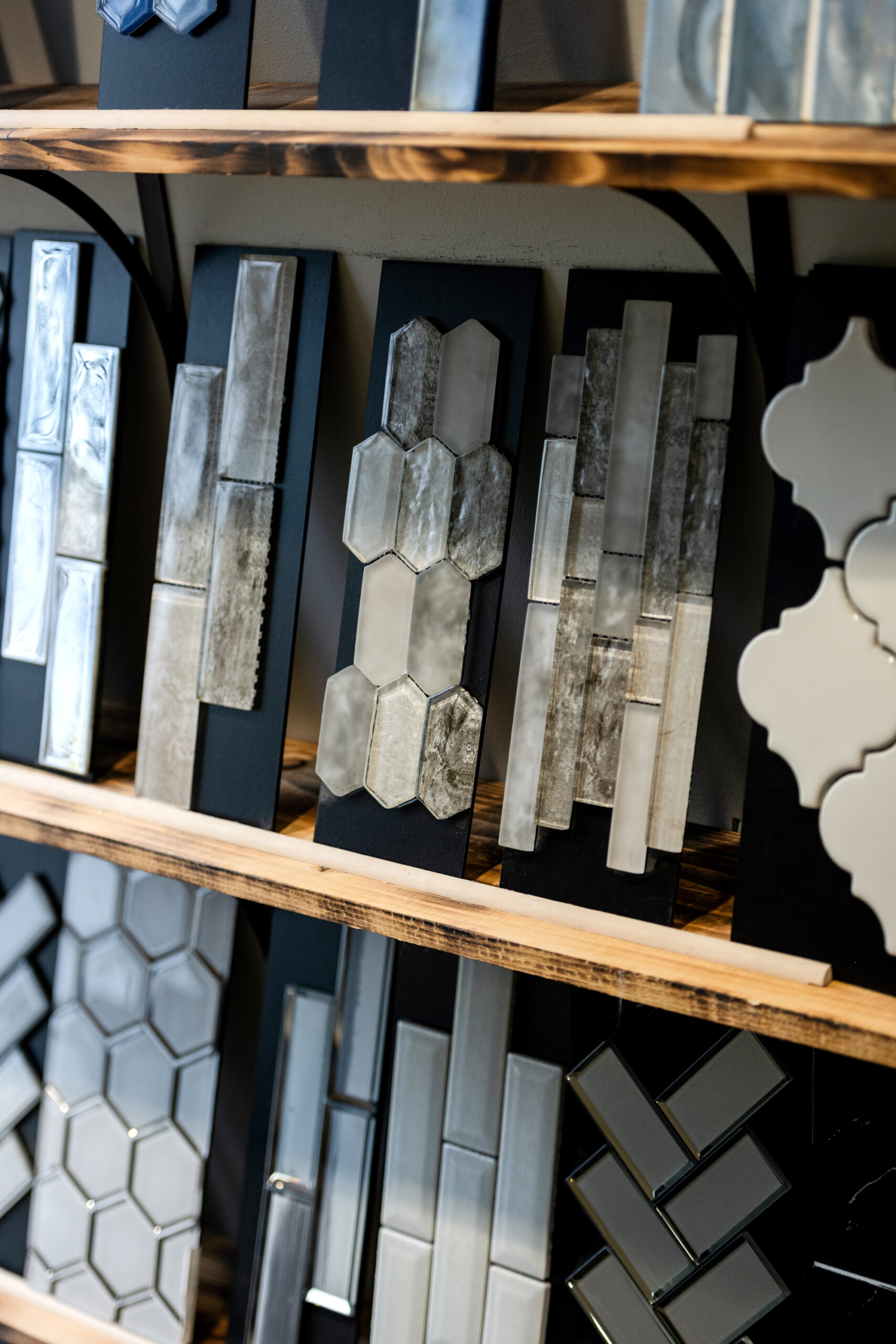 Assorted decorative tiles displayed vertically on wooden shelves, featuring various shapes such as hexagons, rectangles, and quatrefoils in neutral and metallic tones—perfect for any bath remodel or kitchen remodel.