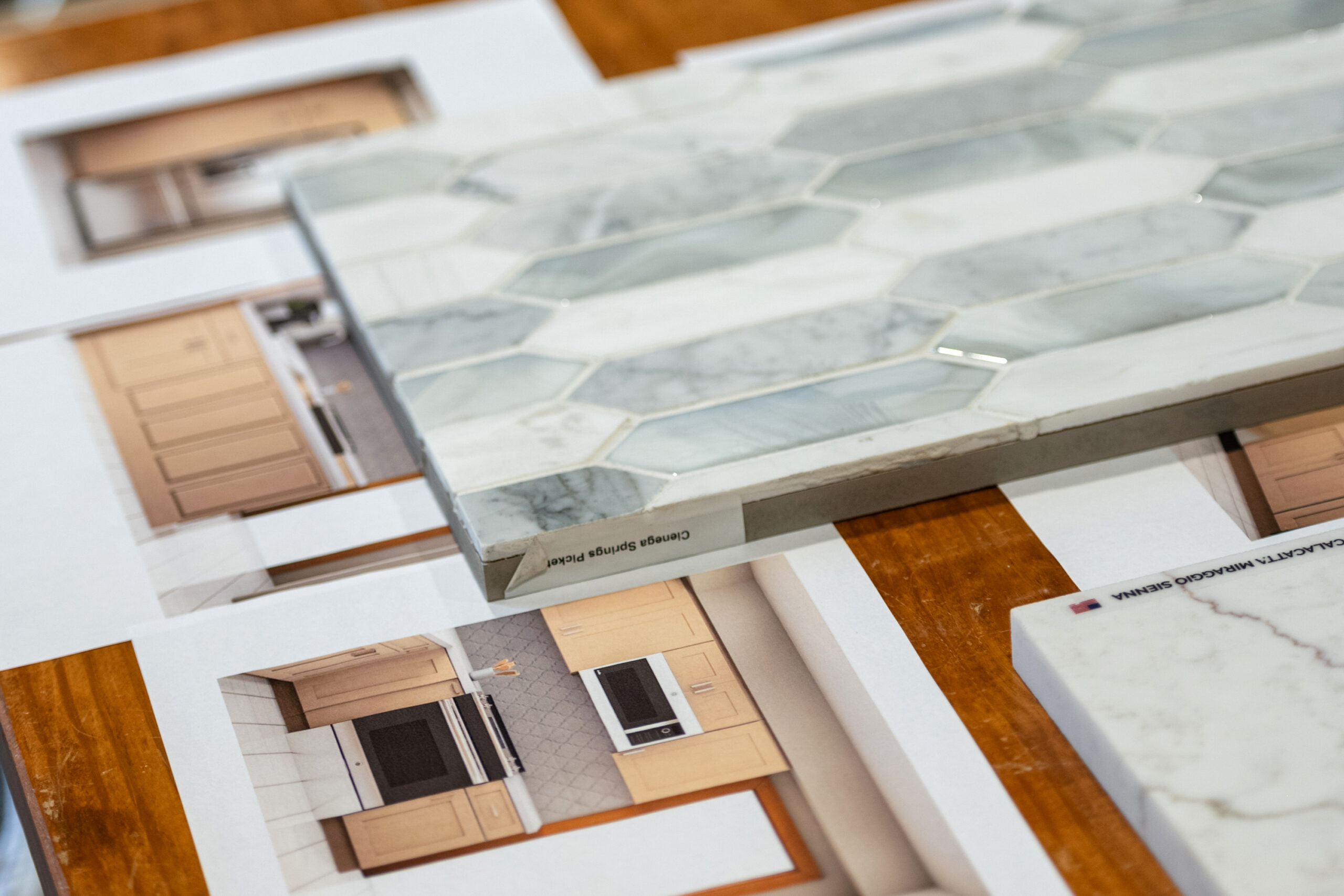 A close-up of a marble hexagon tile sample placed on top of kitchen design renderings with wood cabinets and marble countertops on a wooden table, perfect inspiration for your next kitchen renovation.