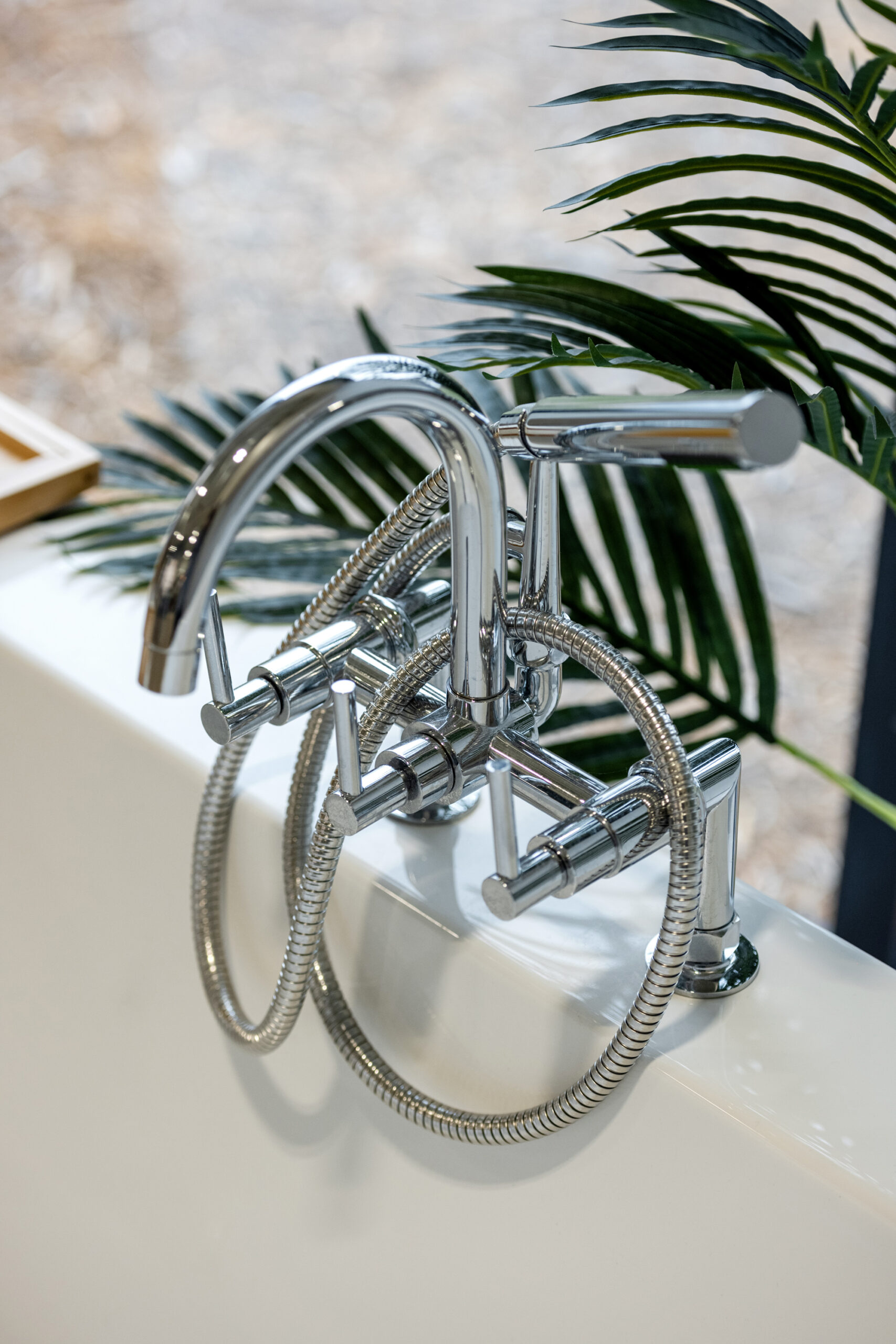 A modern chrome bathtub faucet with a handheld showerhead and flexible hose is mounted on a white tub—a perfect touch for any bath remodel. Green palm leaves are visible in the background.