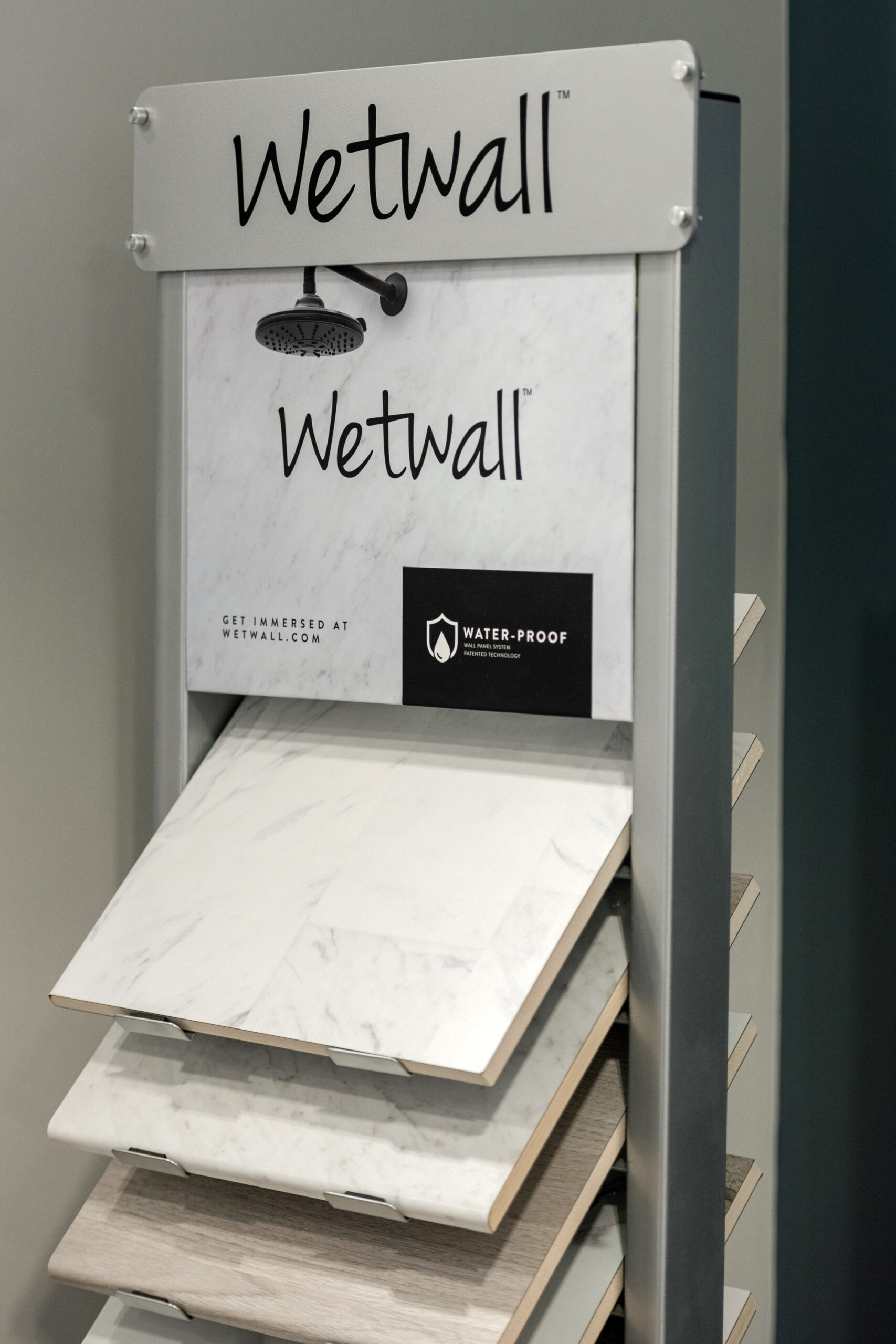 A display stand labeled Wetwall showcases samples of wall panels with marble and wood-like finishes—ideal for your next bath remodel—with a sign and showerhead icon highlighting their waterproof property.
