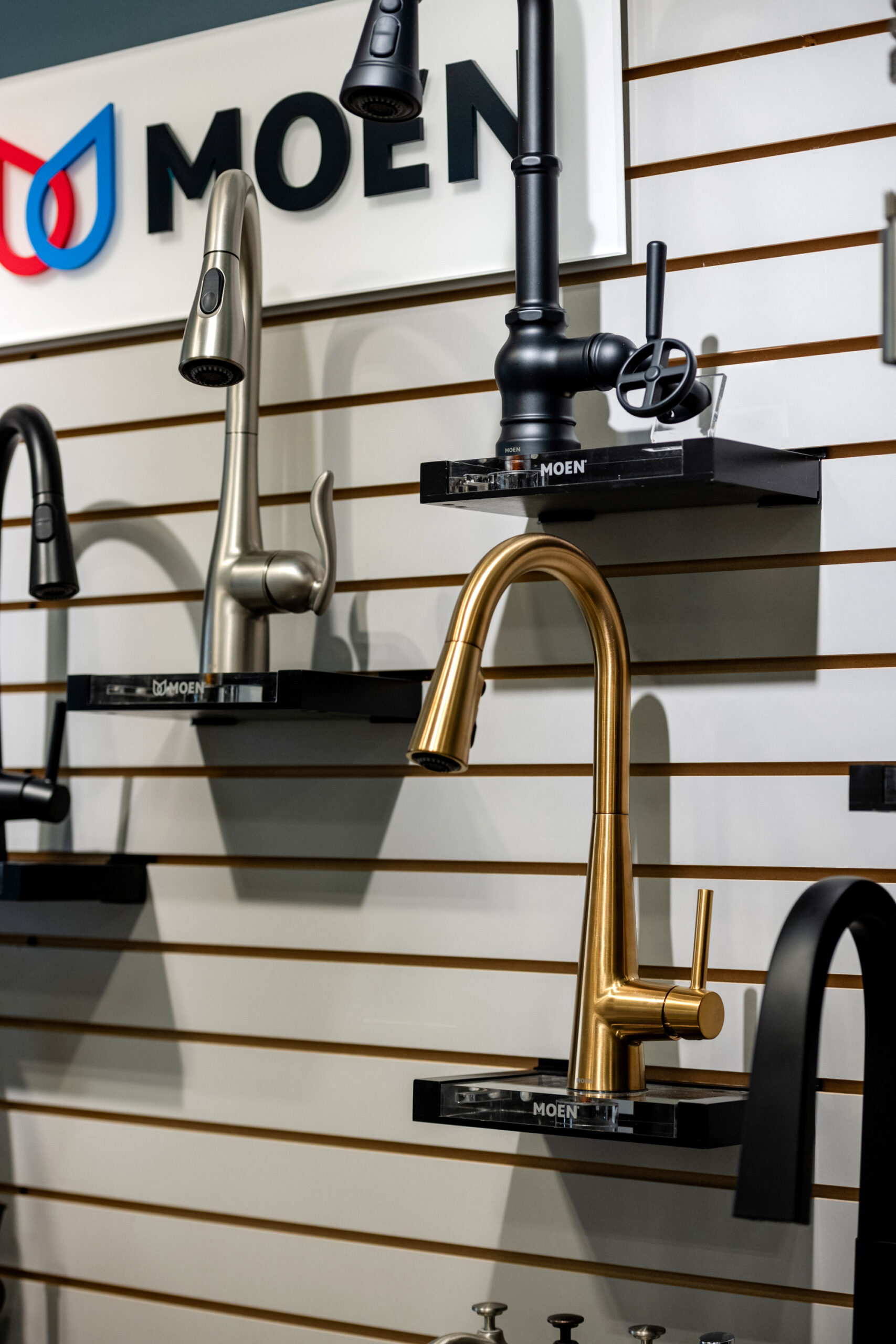 A display wall showcases modern Moen kitchen faucets in matte black, brushed metal, and gold finishes—perfect inspiration for your next kitchen remodel—mounted on black shelves beneath a Moen logo sign.