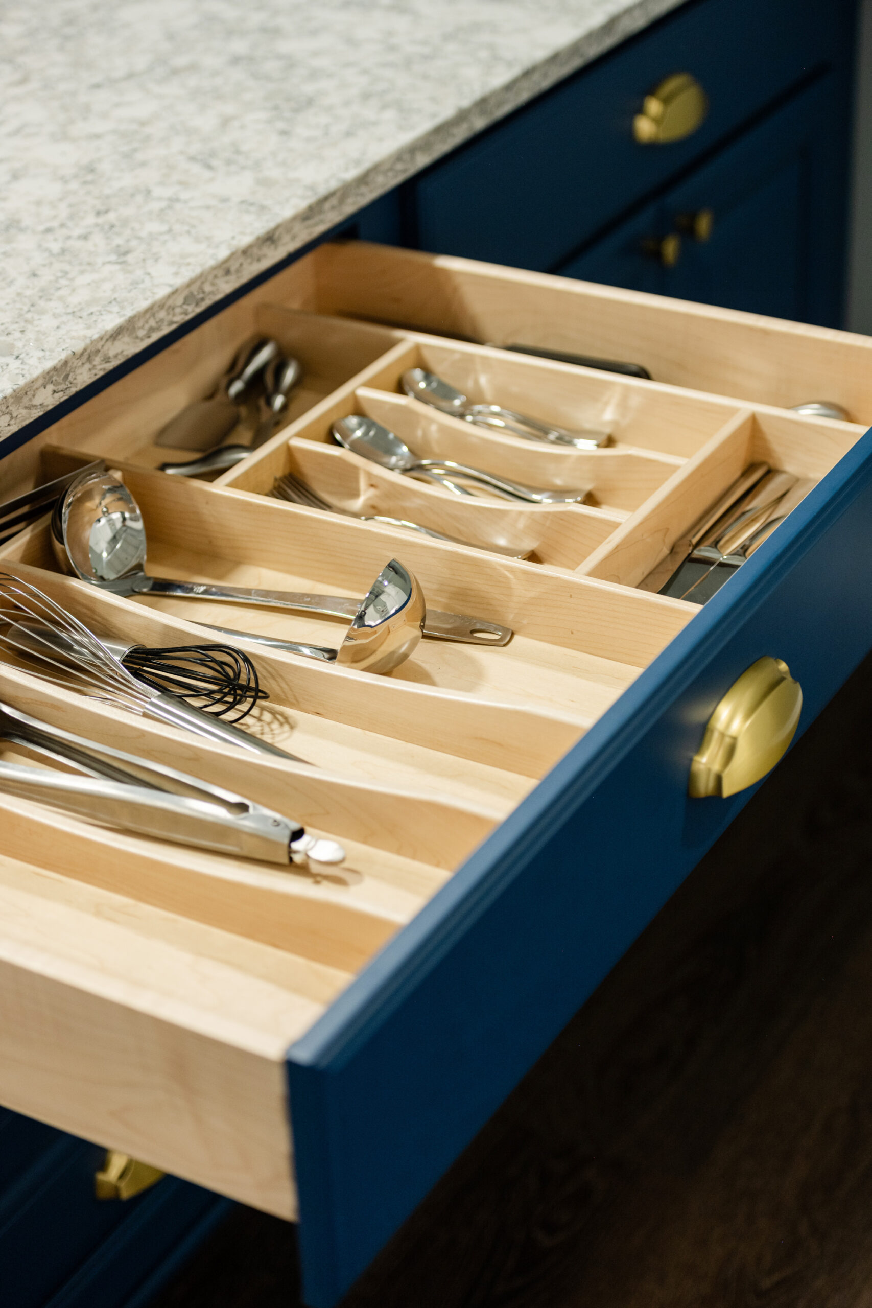 An open kitchen drawer with organized sections holds ladles, spoons, and a whisk in a blue cabinet with gold handles under a speckled countertop—perfect inspiration for your next kitchen remodel.