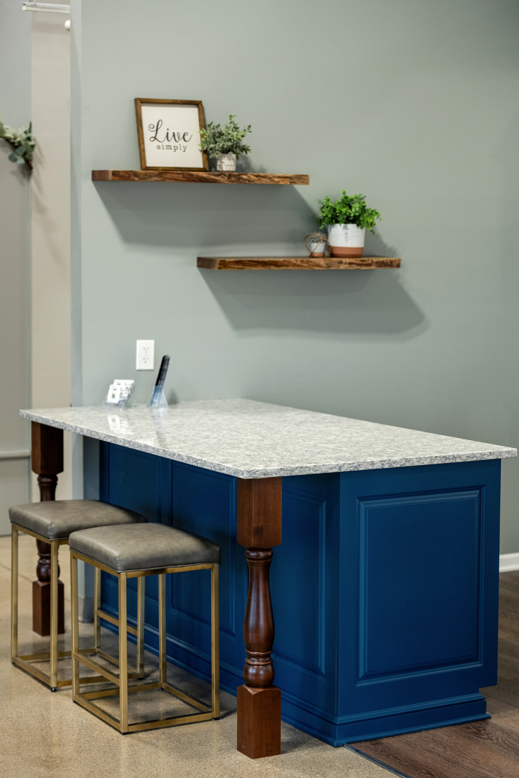 A modern kitchen corner features a blue island with a white marble countertop, two cushioned stools, and wooden shelves on a gray wall decorated with potted plants and a framed Live simply sign by a top kitchen remodeling company Columbus Ohio.