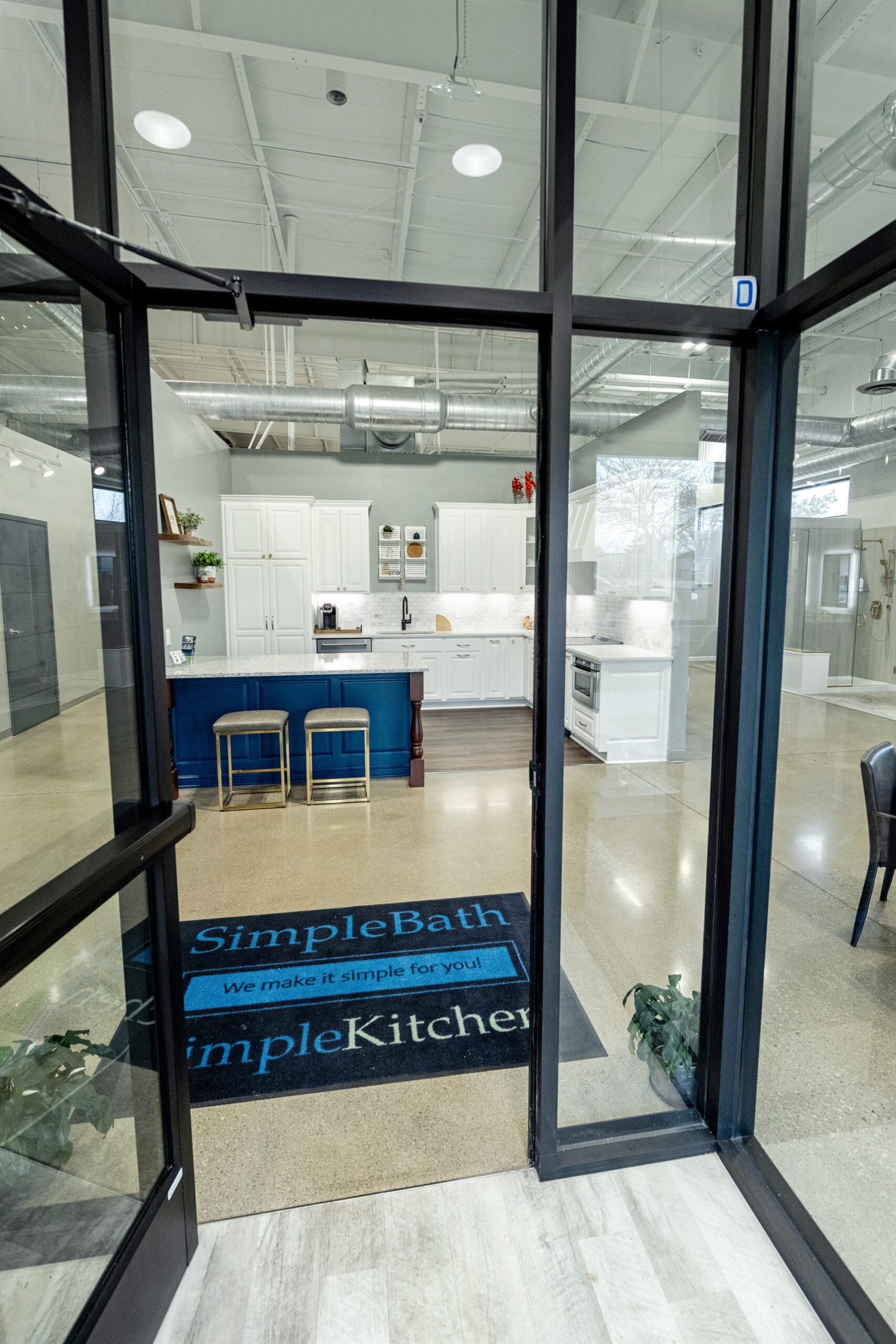 View through a glass door into a modern showroom by a top kitchen remodeling company Columbus Ohio, featuring a white kitchen with a blue island, two stools, and “SimpleBath SimpleKitchens” branded mats on a polished floor.