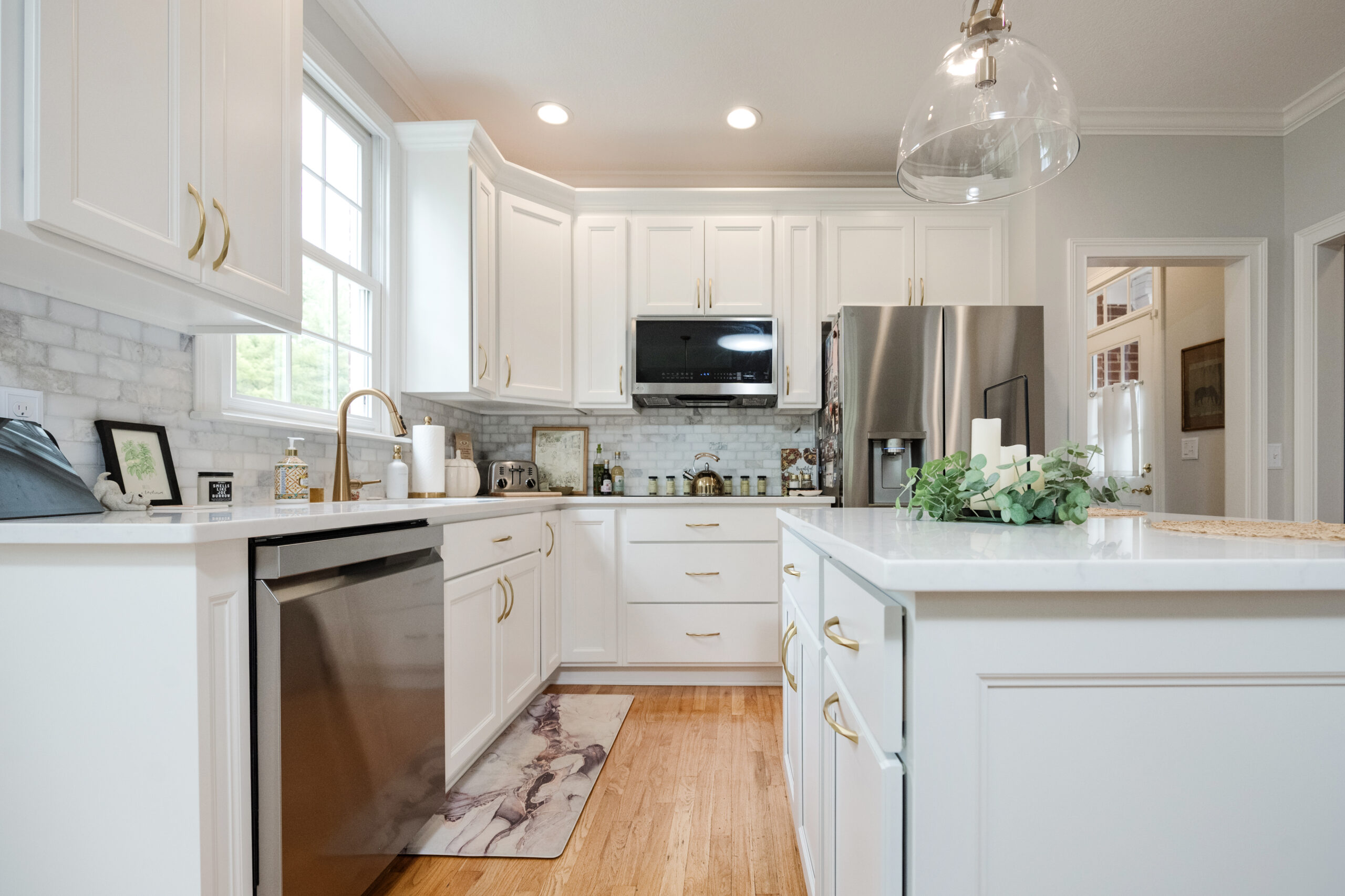 A bright, modern kitchen with white cabinets, gold handles, stainless steel appliances, a wooden floor, and a large island—beautifully designed by a leading kitchen remodeling company in Columbus, Ohio—with sunlight streaming through the window.