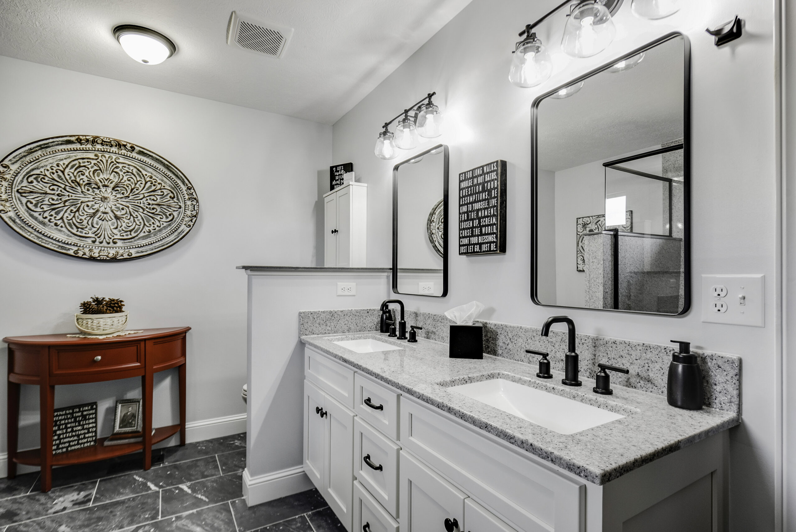 Modern bathroom with double sinks, granite countertop, black fixtures, two mirrors, wall lights, and a red accent table under a decorative wall piece—showcasing bathroom renovation ideas from a top bath remodel expert. Gray tile floor and white cabinetry.