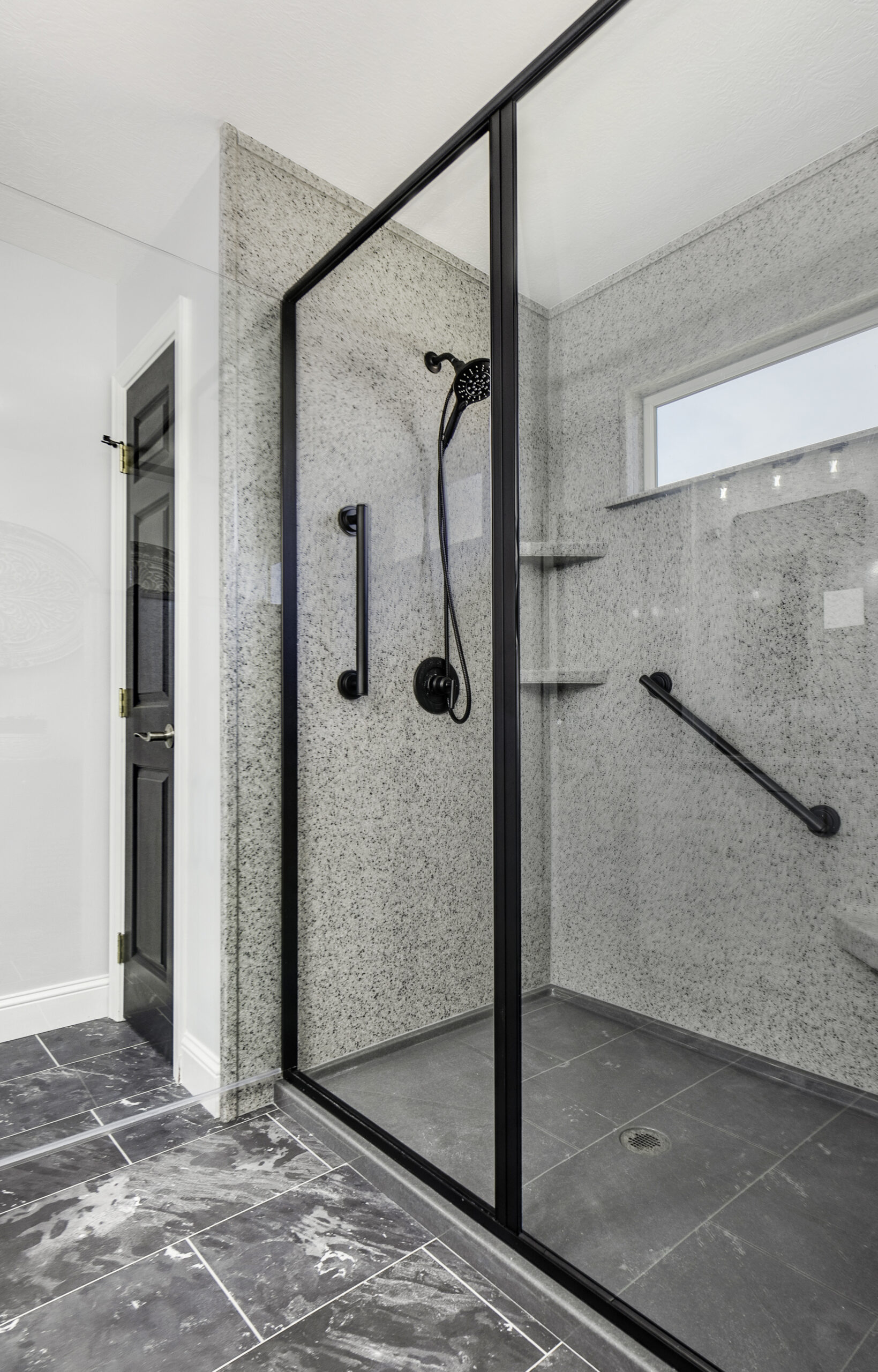 Modern bathroom by a leading bathroom remodeling company in Columbus, Ohio, with a spacious walk-in shower enclosed by glass panels, black fixtures, built-in shelves, gray textured wall tiles, and dark gray marble flooring.