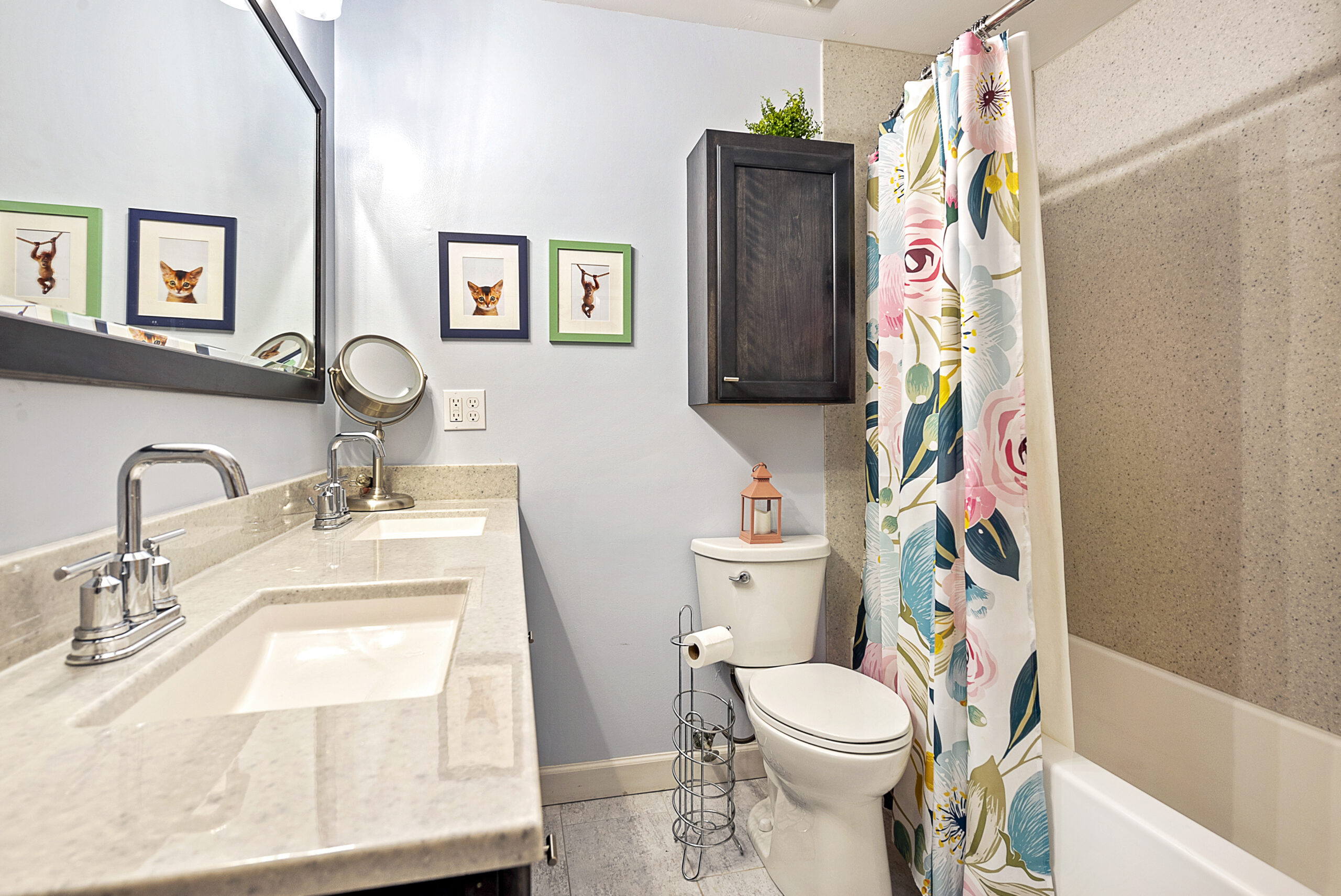 A bright bathroom with a floral shower curtain, dual sinks on a light countertop, wall-mounted cabinet, and three animal prints on a pale blue wall—styled to complement any kitchen remodel or renovation. A lantern sits atop the toilet tank.