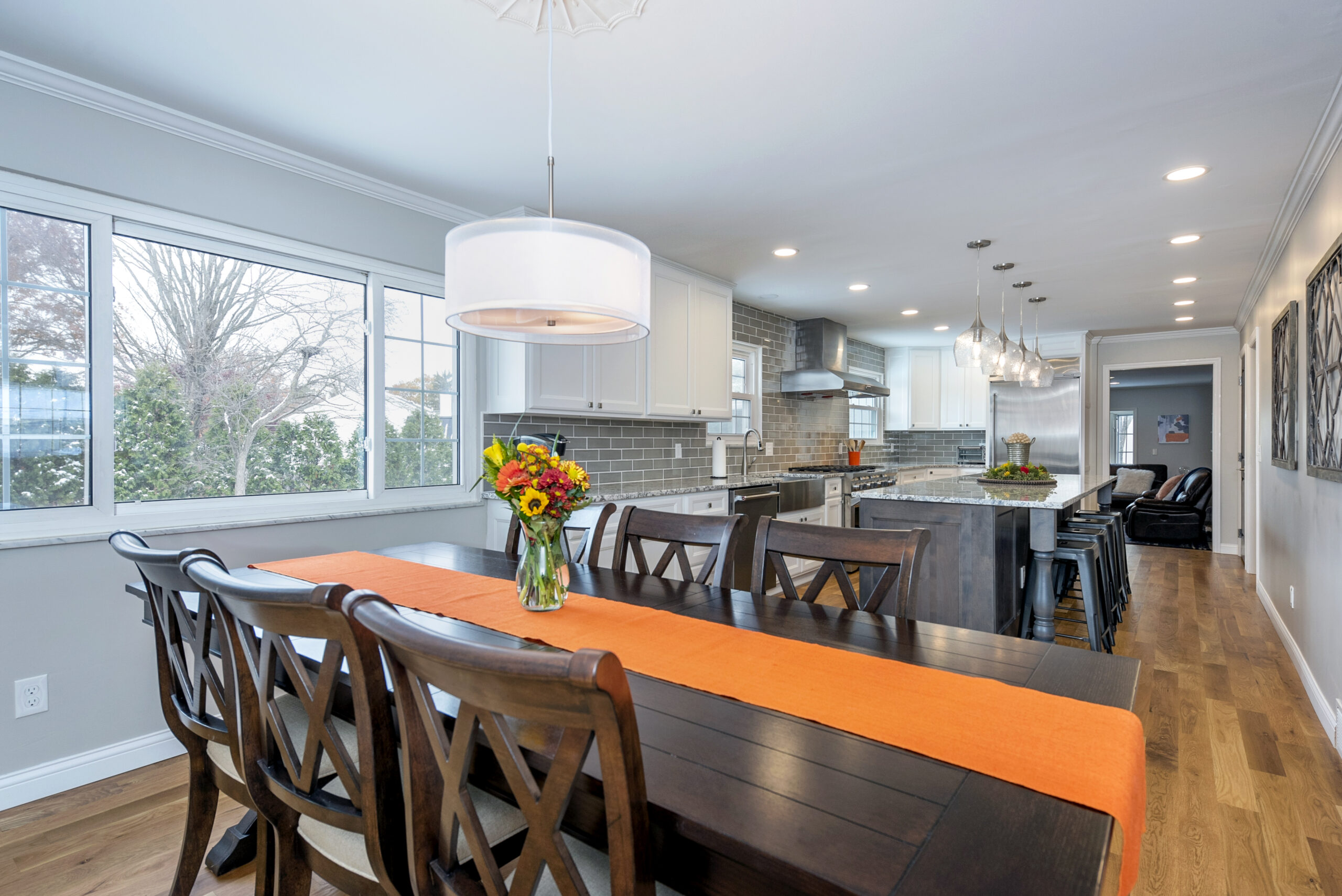 A modern kitchen and dining area with a wooden table, dark chairs, an orange runner, and a vase of flowers. Large windows, pendant lights, and gray cabinets complete the bright space by a leading kitchen remodeling company Columbus Ohio.