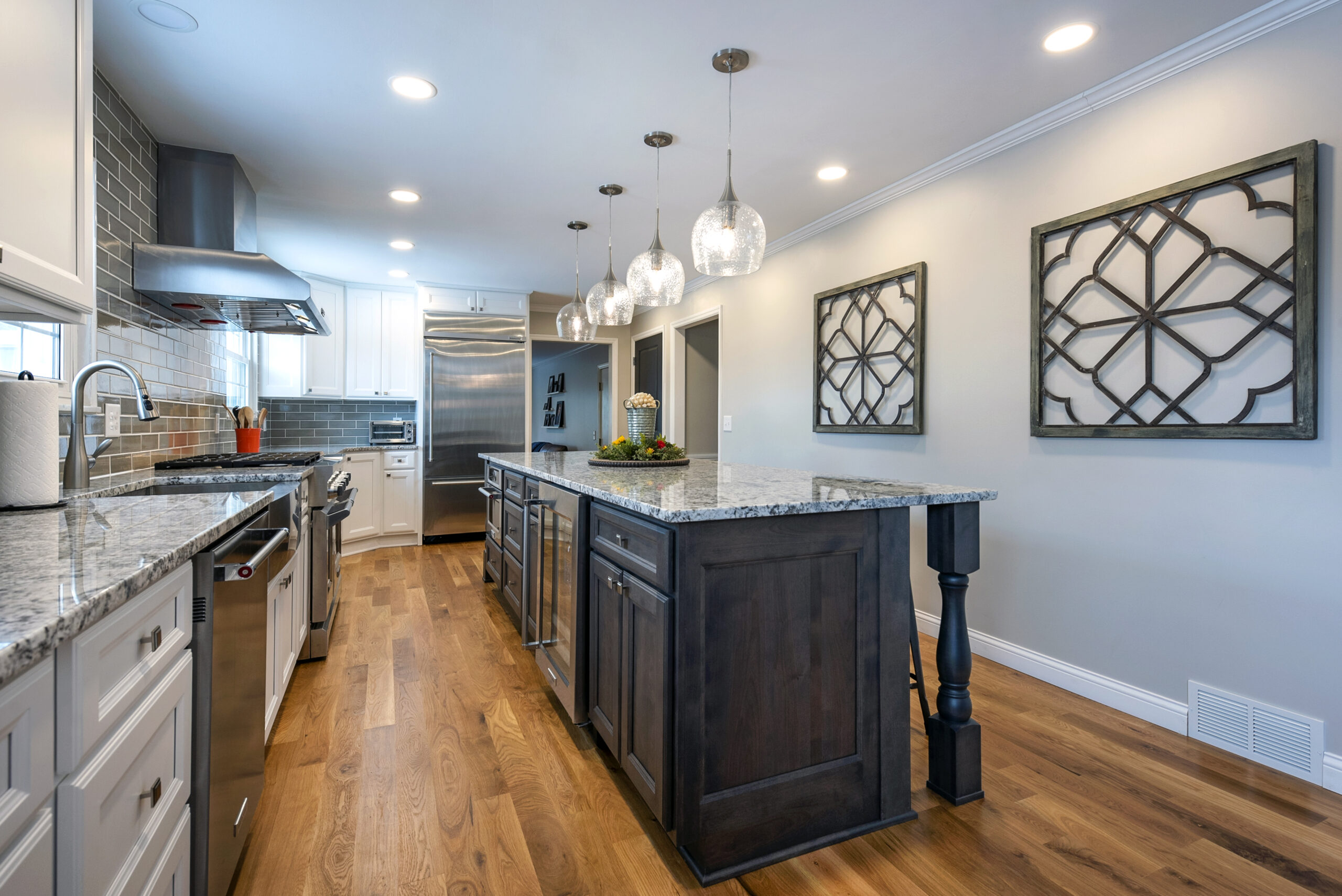 Modern kitchen with wooden floors, a large island with granite countertop, stainless steel appliances, and pendant lights above the island—perfect inspiration for your next kitchen renovation. White cabinets and gray backsplash complete the space.