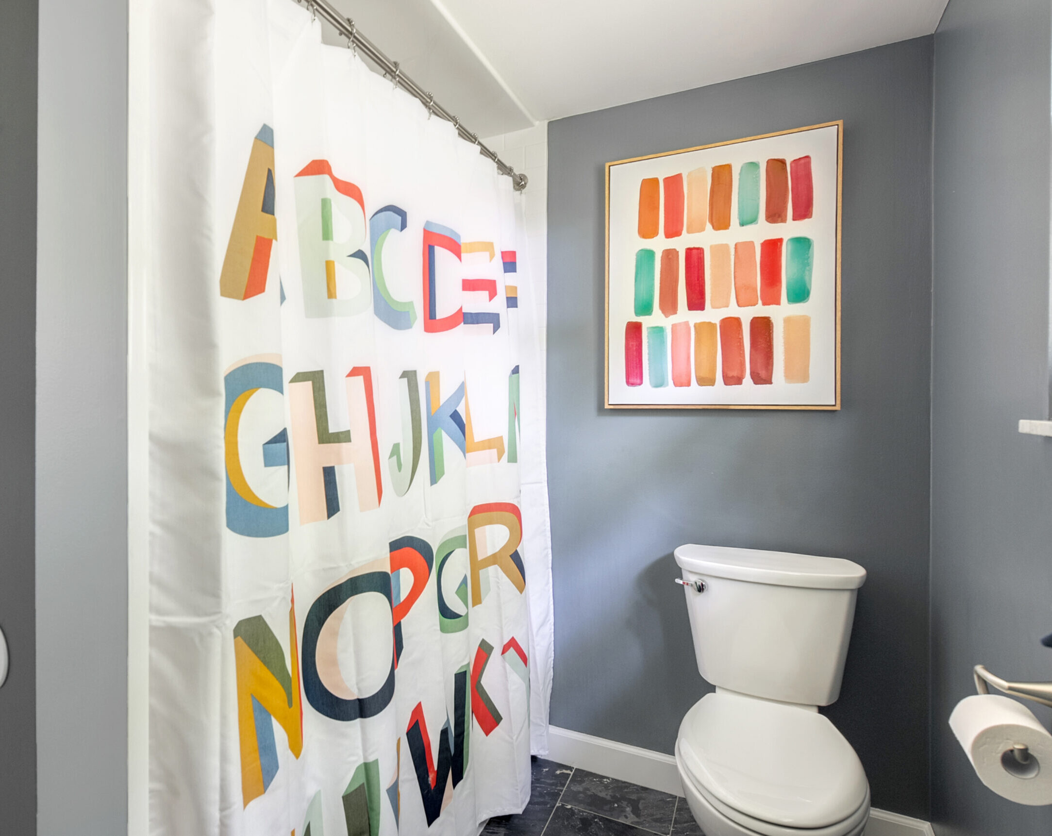 A bath remodel features a colorful alphabet-patterned shower curtain, a white toilet, gray walls, and a framed abstract painting with red, orange, yellow, and green brushstrokes above the toilet.