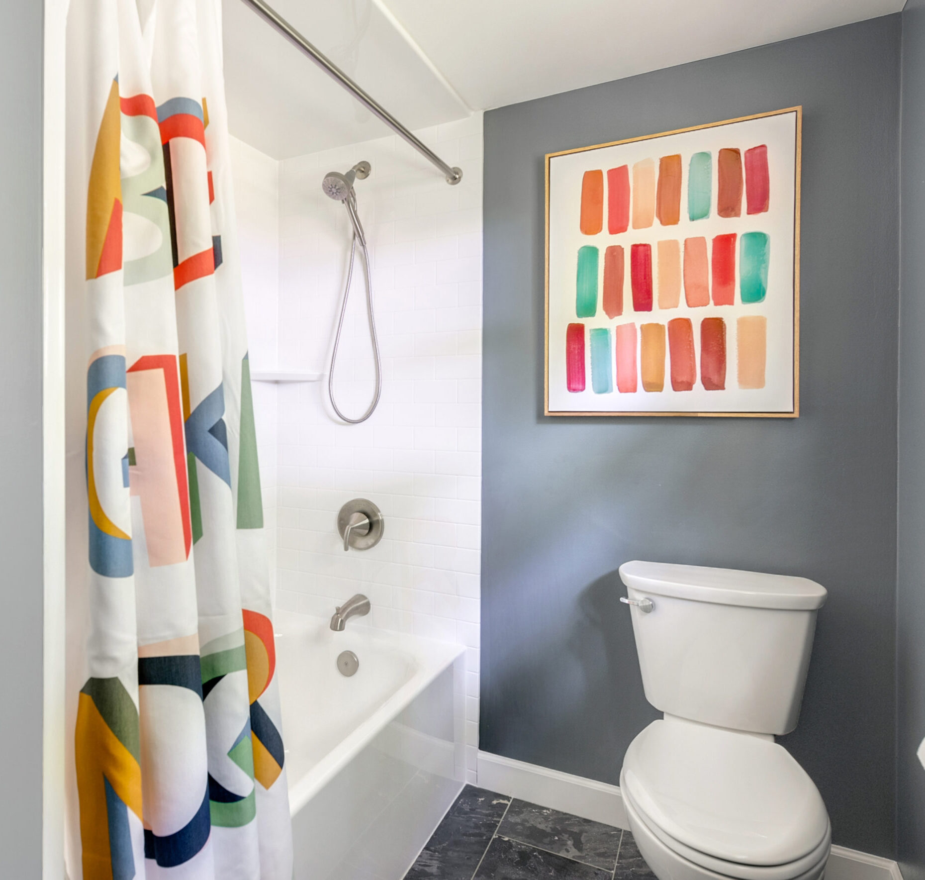 Modern bathroom renovation with gray walls, white bathtub and toilet, colorful abstract art, and a shower curtain featuring large, multicolored letters. Dark tile flooring completes this stylish bath remodel.
