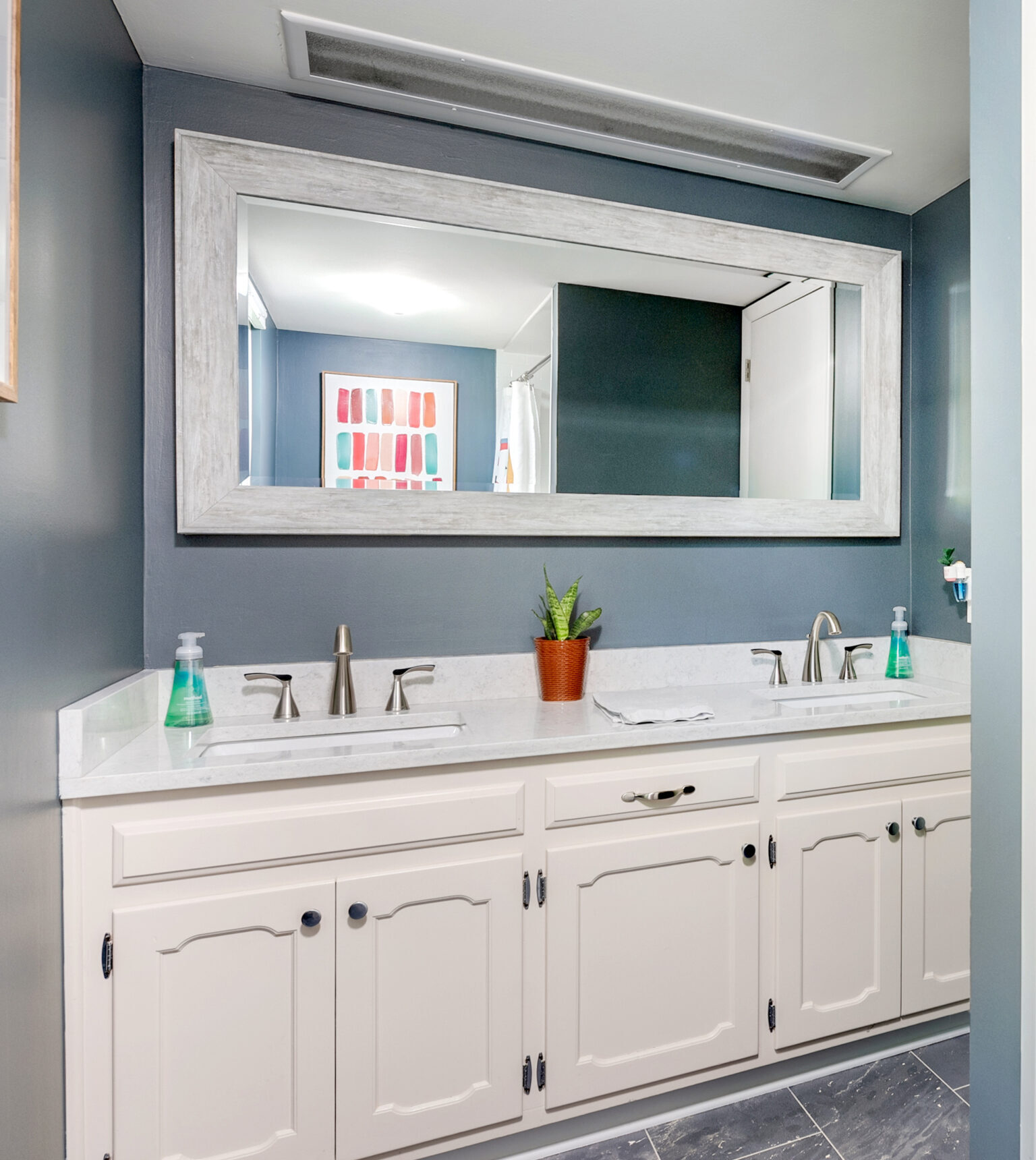 Double bathroom vanity with white cabinets, marble countertop, two sinks, and silver faucets is perfect for your bathroom renovation. A large framed mirror reflects a potted plant and soap dispensers; the walls are painted deep blue-gray.