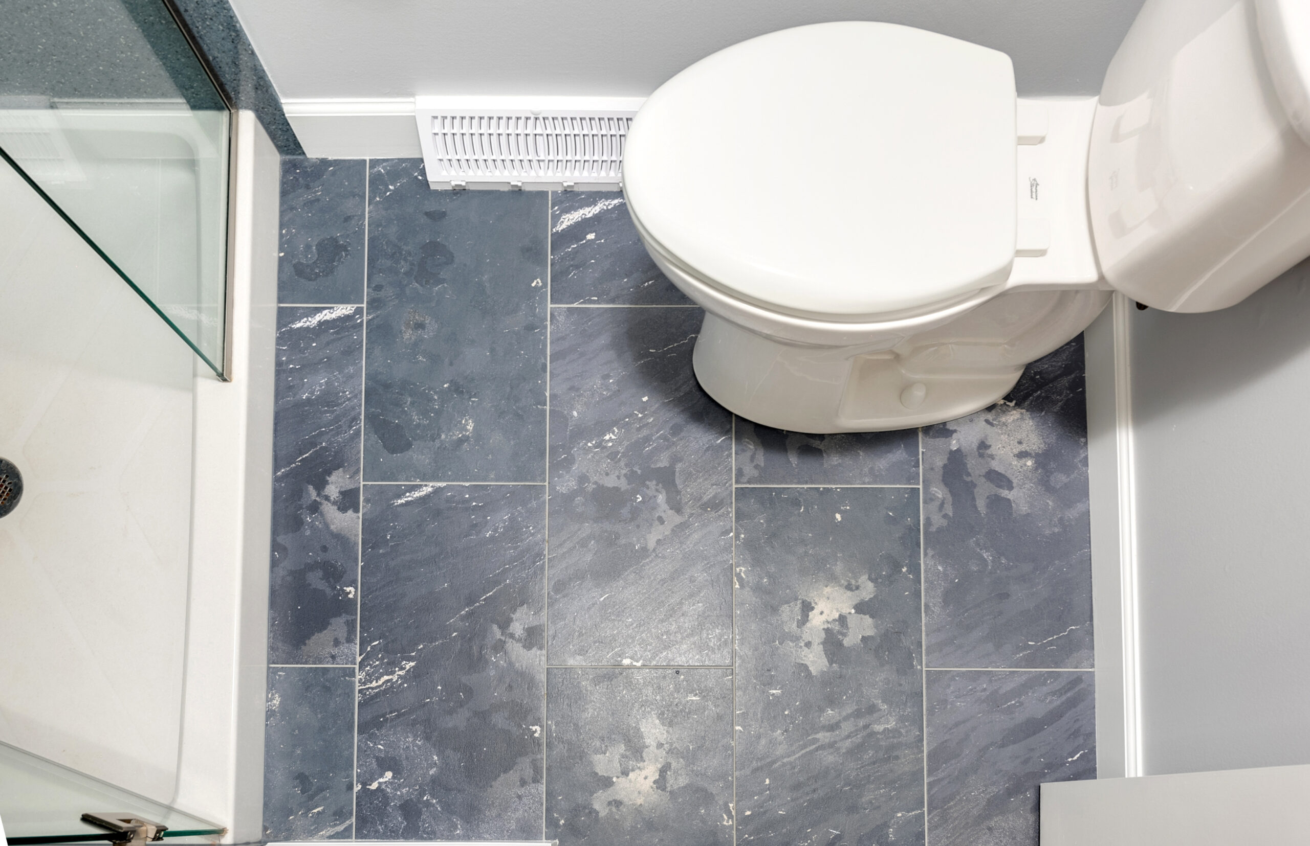 Overhead view of a bathroom corner showcases a white toilet on grayish-blue, marbled tile—ideal inspiration for your next bathroom renovation by a top bathroom remodeling company Columbus Ohio homeowners trust. Glass shower and floor vent also visible.