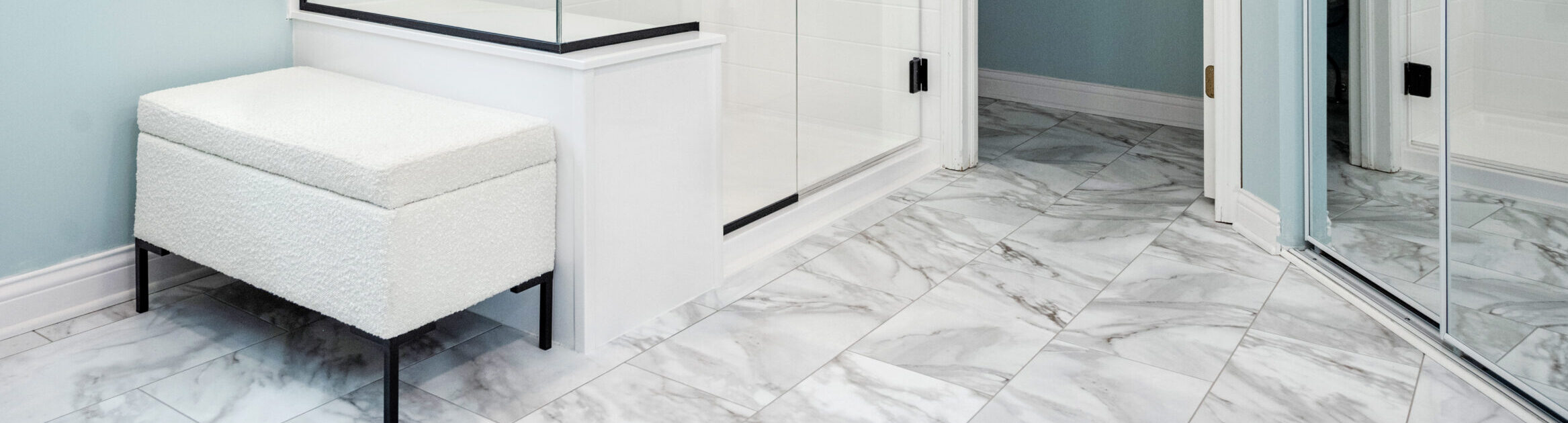 Modern bathroom with gray marble tile floor, a glass shower enclosure, a light upholstered bench, and light blue walls. A large mirror reflects part of the room—perfect inspiration for your next bathroom renovation.