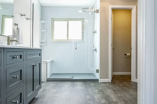 Modern bathroom renovation by a leading bathroom remodeling company in Columbus, Ohio, featuring a gray vanity, large glass-enclosed shower with built-in bench, wood-style flooring, and a separate toilet room visible through an open door.