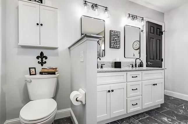 Modern bathroom renovation with white cabinets, double vanity, and two mirrors. Toilet with shelving and decor above sits next to a partial privacy wall. Black fixtures and accents complement the gray walls and marble floor tiles.