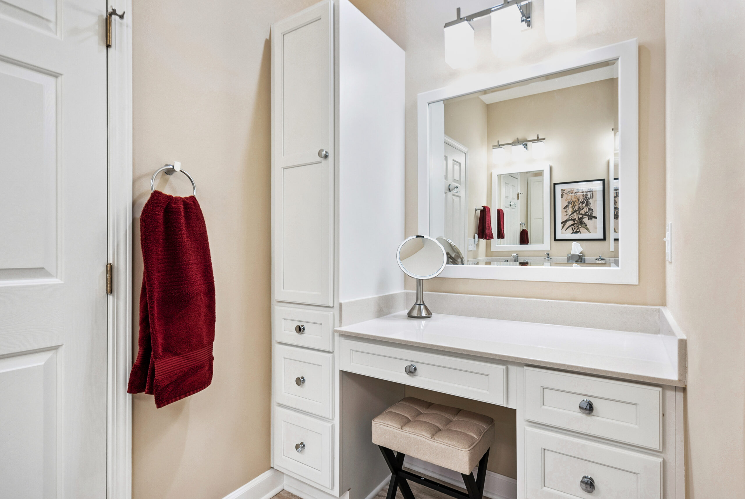 A white bathroom vanity with drawers, a large mirror, a small round tabletop mirror, a padded stool, and two red towels hanging on a silver ring—all against beige walls—showcase the style you can expect from a trusted bath remodel by a leading bathroom remodeling company in Columbus, Ohio.