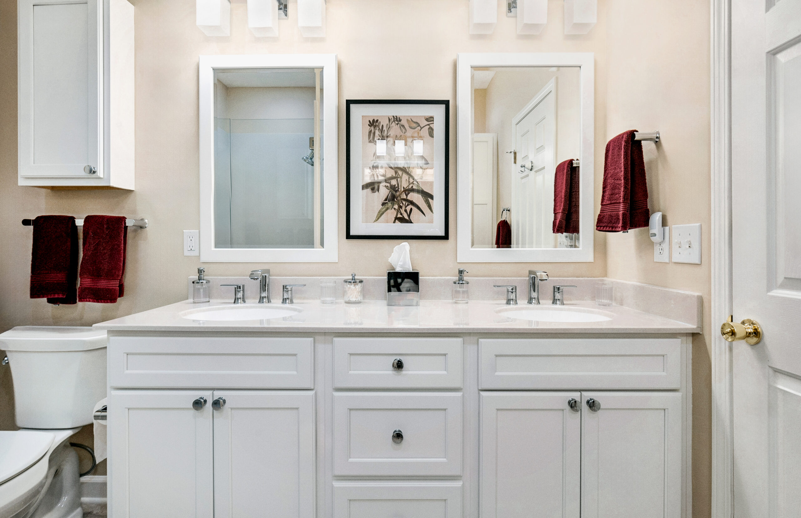 A modern bathroom renovation features a double sink vanity, white cabinets, two mirrors, red towels, a toilet, and a framed floral picture. Beige walls and a door with a gold handle complete this stylish bath remodel.