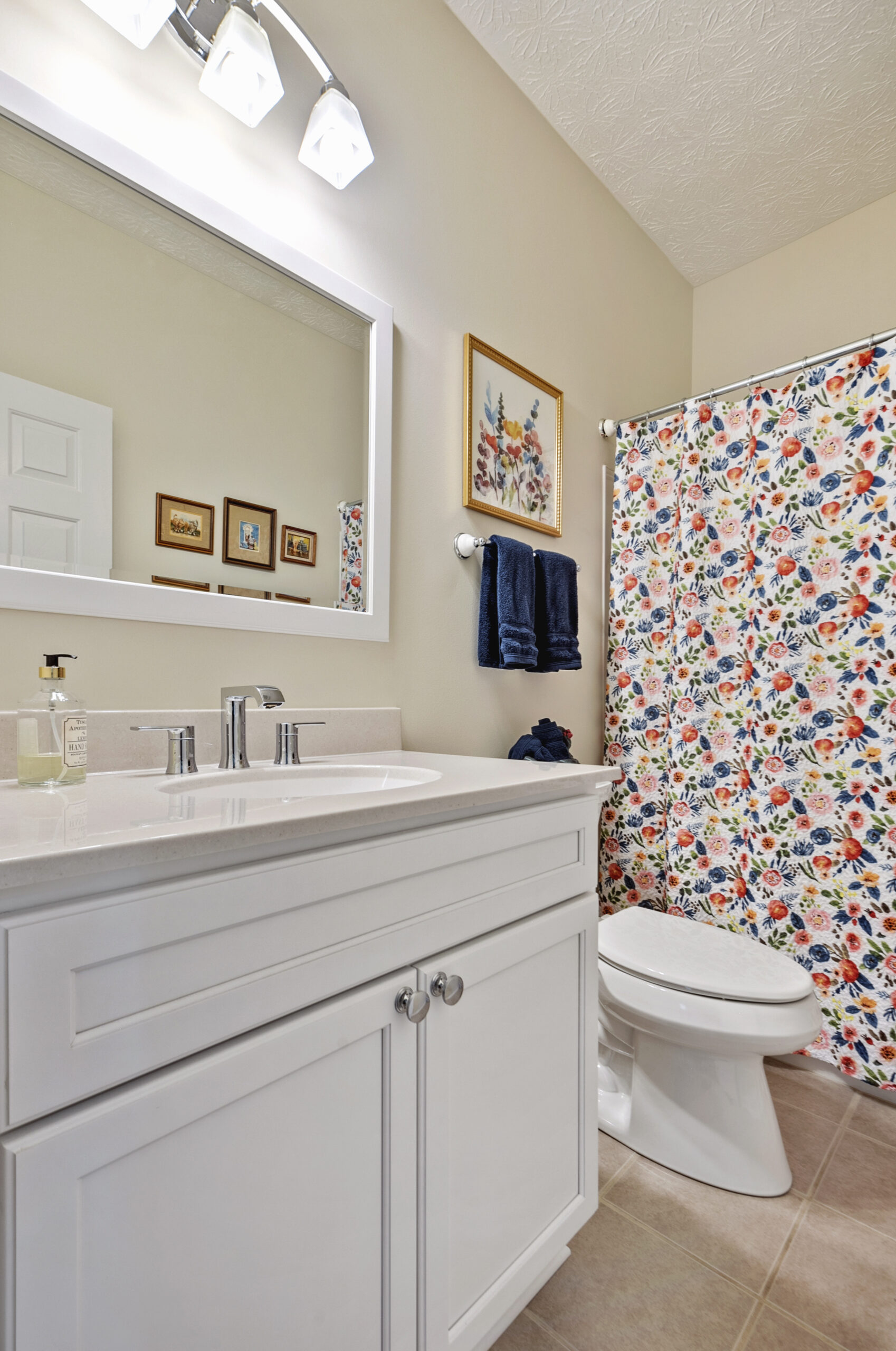 A bright bathroom renovation features a white vanity, silver faucet, and mirror above. There’s a toilet and a shower with a colorful floral curtain. Blue towels hang on a rack, and framed art decorates the beige walls.