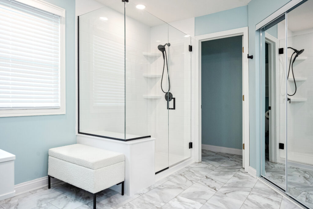 Modern bathroom renovation with a glass-enclosed shower, black fixtures, white tile walls, marble-patterned floor, cushioned bench, light blue walls, and a large mirror—perfect inspiration from a top bathroom remodeling company in Columbus Ohio.