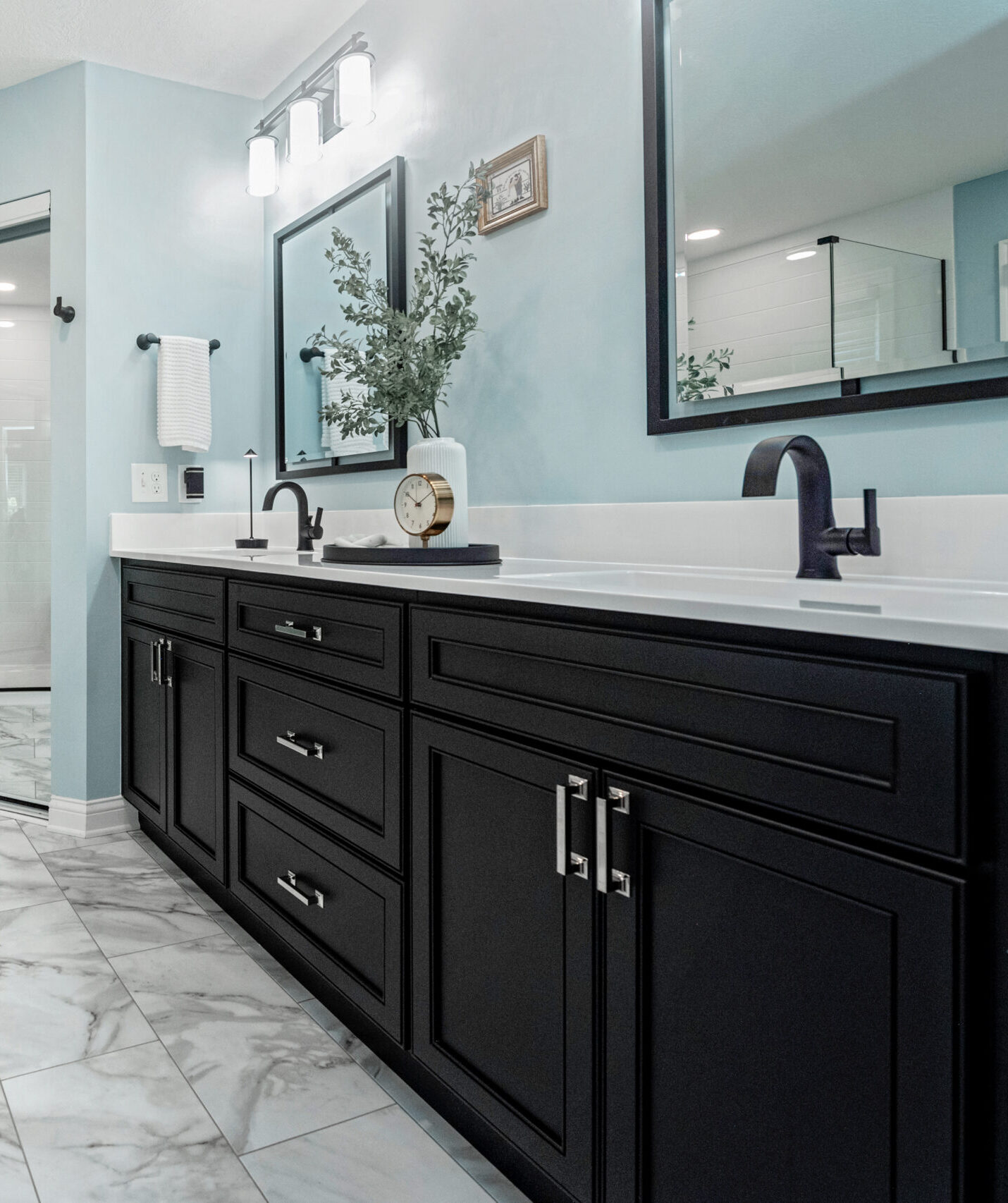 Modern bathroom with a long black vanity, white countertop, black faucets, and a large mirror. Perfect for a bath remodel, the light blue walls and marble tiles create a fresh look, while a plant and clock add style to the counter.