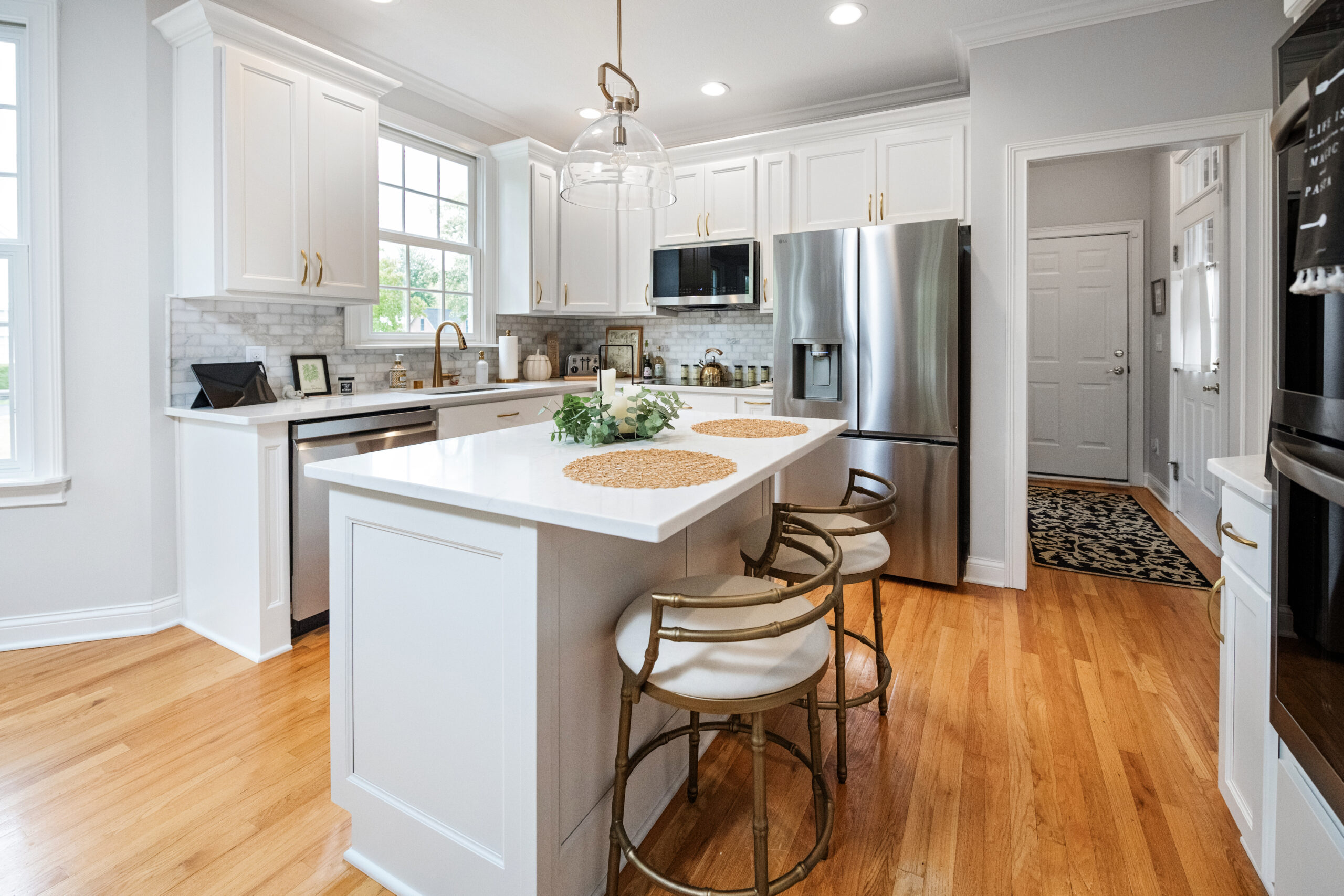 Bright, modern kitchen with white cabinets, stainless steel appliances, a central island with two stools, pendant lighting, wood floors, and large windows—expertly crafted by a leading kitchen remodeling company Columbus Ohio.