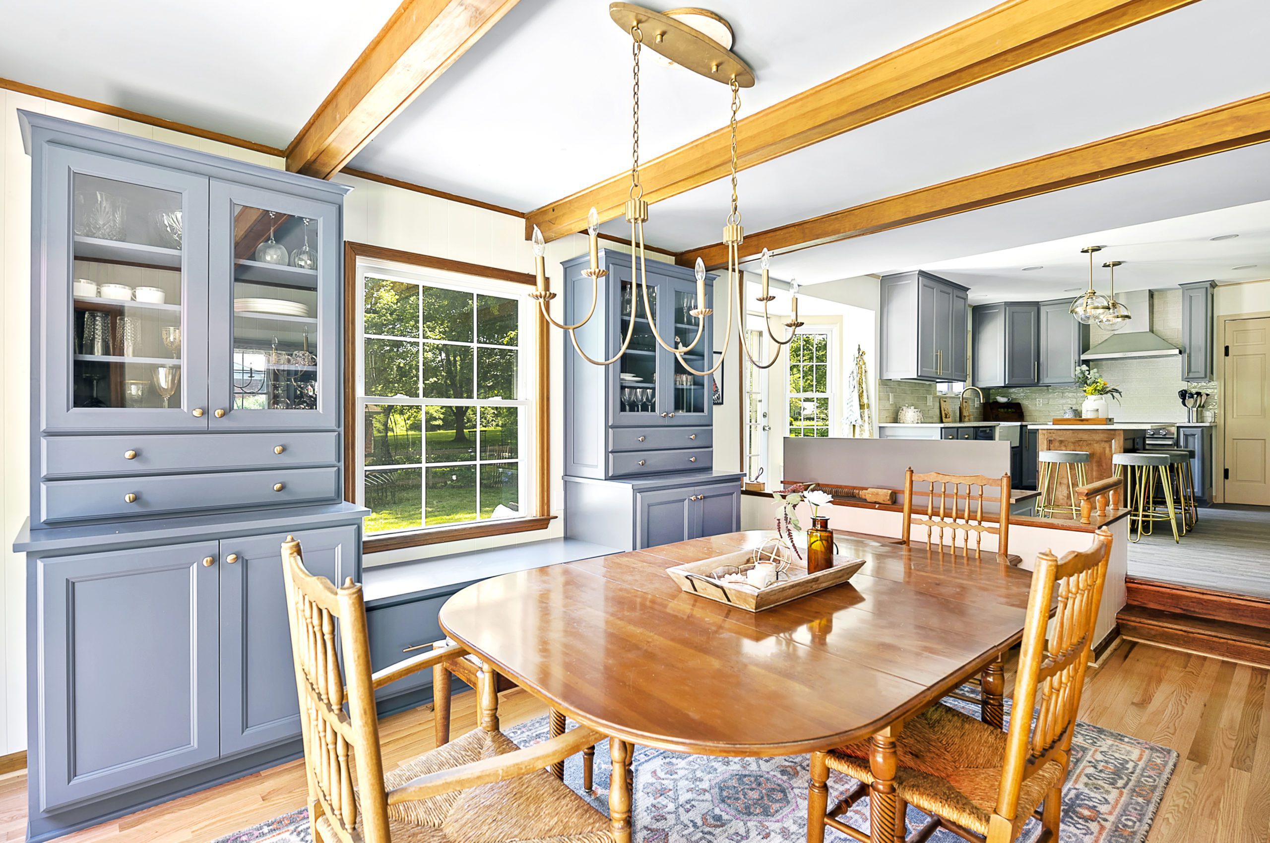 Bright dining room with large windows, a wooden table and chairs, blue cabinetry, a chandelier, and a view into a modern kitchen remodel featuring gray cabinets and a breakfast bar. Natural light fills the space.