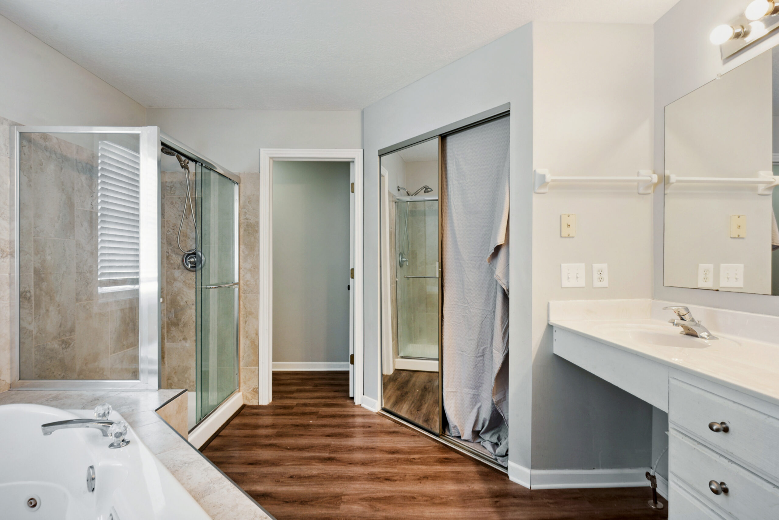Modern bathroom renovation with a jetted tub, glass-enclosed shower, double vanity with large mirror, and wood-style flooring. A closet with mirrored sliding doors contains another shower with a curtain.