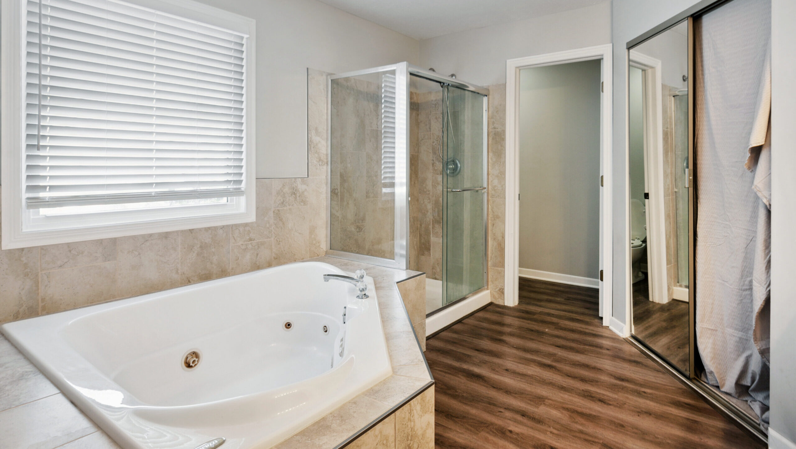 Modern bathroom with a large white bathtub, glass-enclosed shower, tiled walls, wood-style flooring, window with blinds, and mirrored closet door—a perfect example of a stylish bath remodel.
