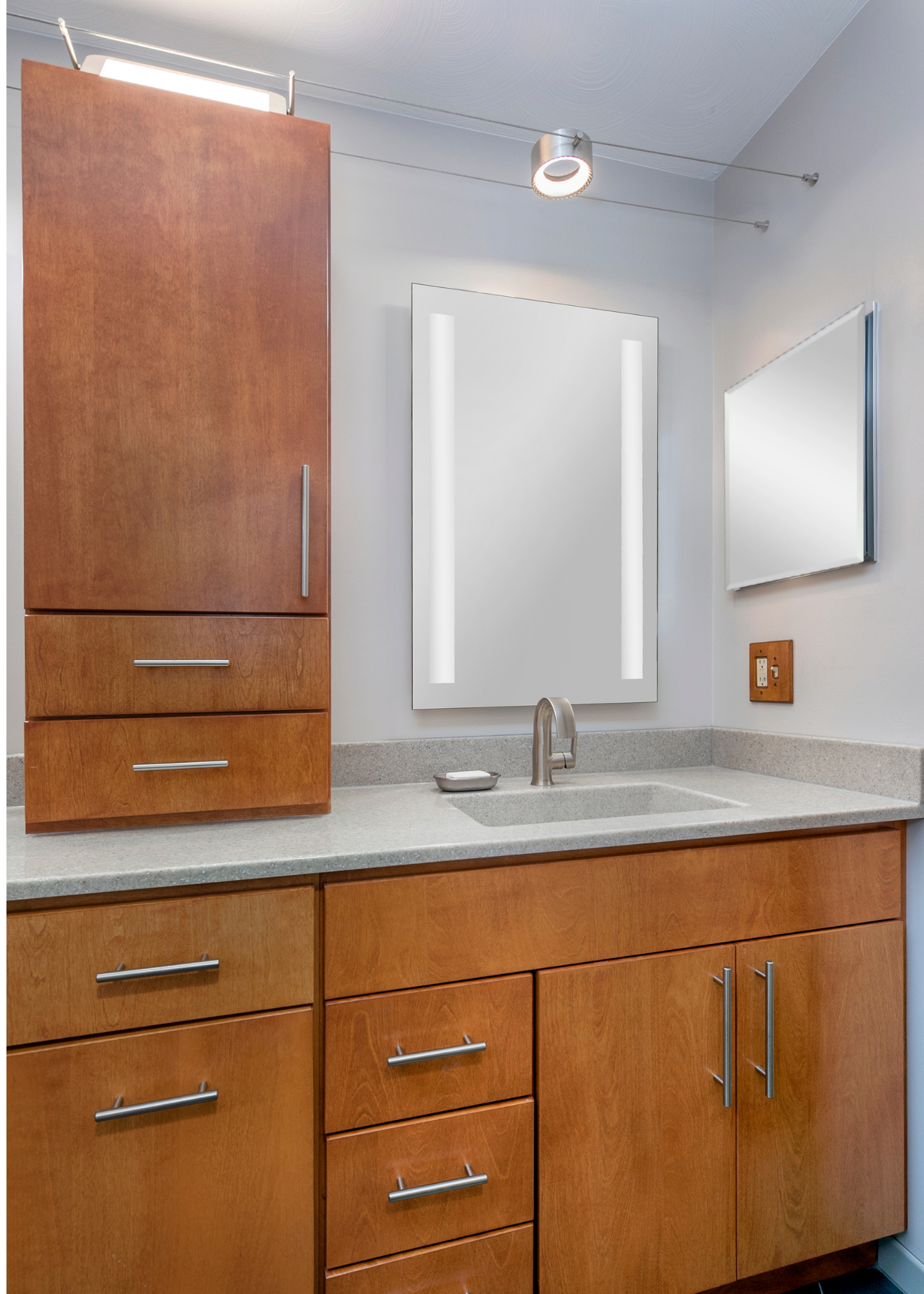 Modern bathroom vanity with light wood cabinets, gray countertop, under-mount sink, large mirror with vertical lights, and a small angled mirror on the wall. Overhead lighting brightens the clean, contemporary space.