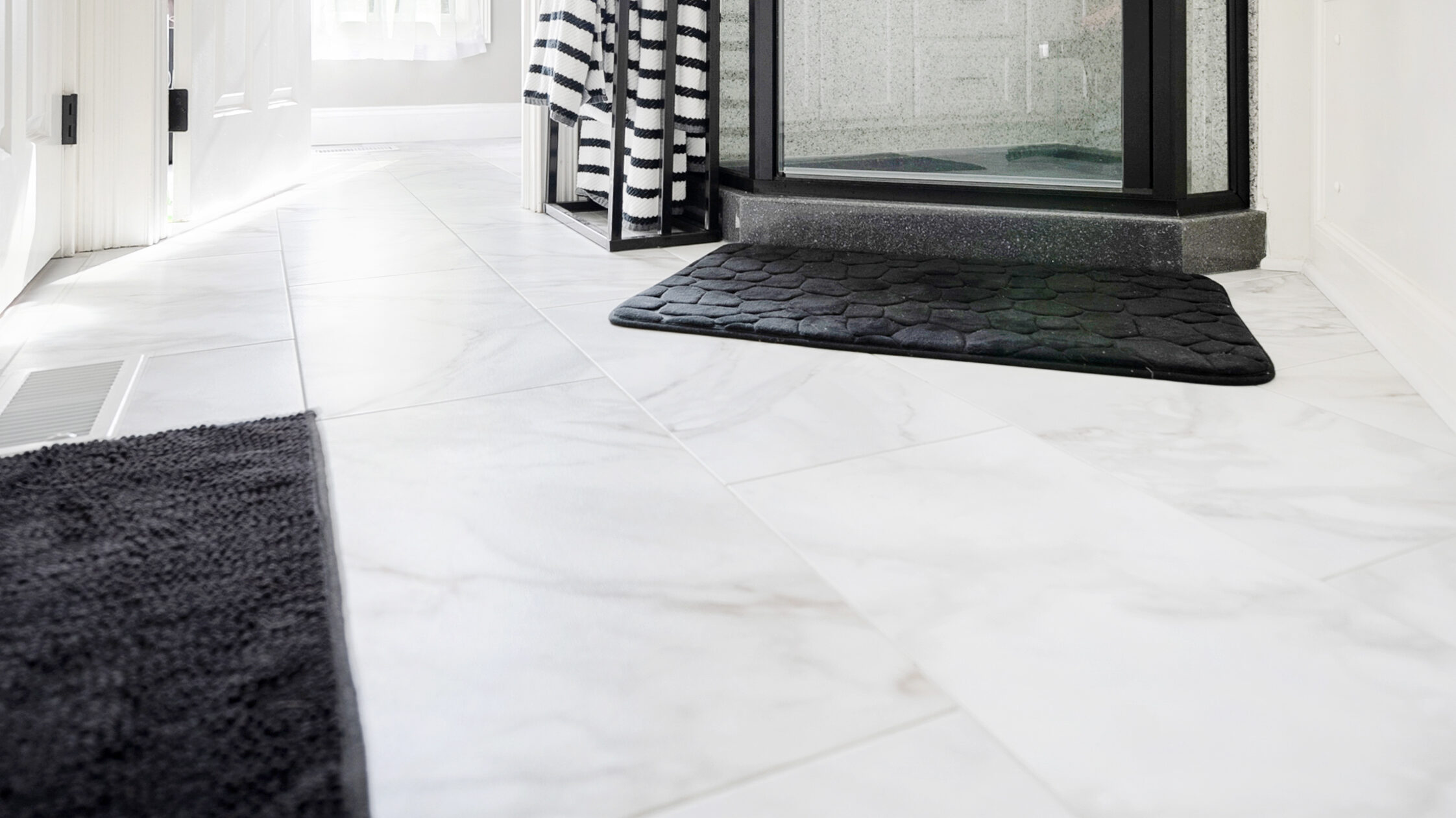 Modern bathroom with a glass shower enclosure, black fixtures, striped towels, white marble floor, black bath mats, and natural light. Expertly designed by a leading bathroom remodeling company Columbus Ohio trusts for quality renovations.
