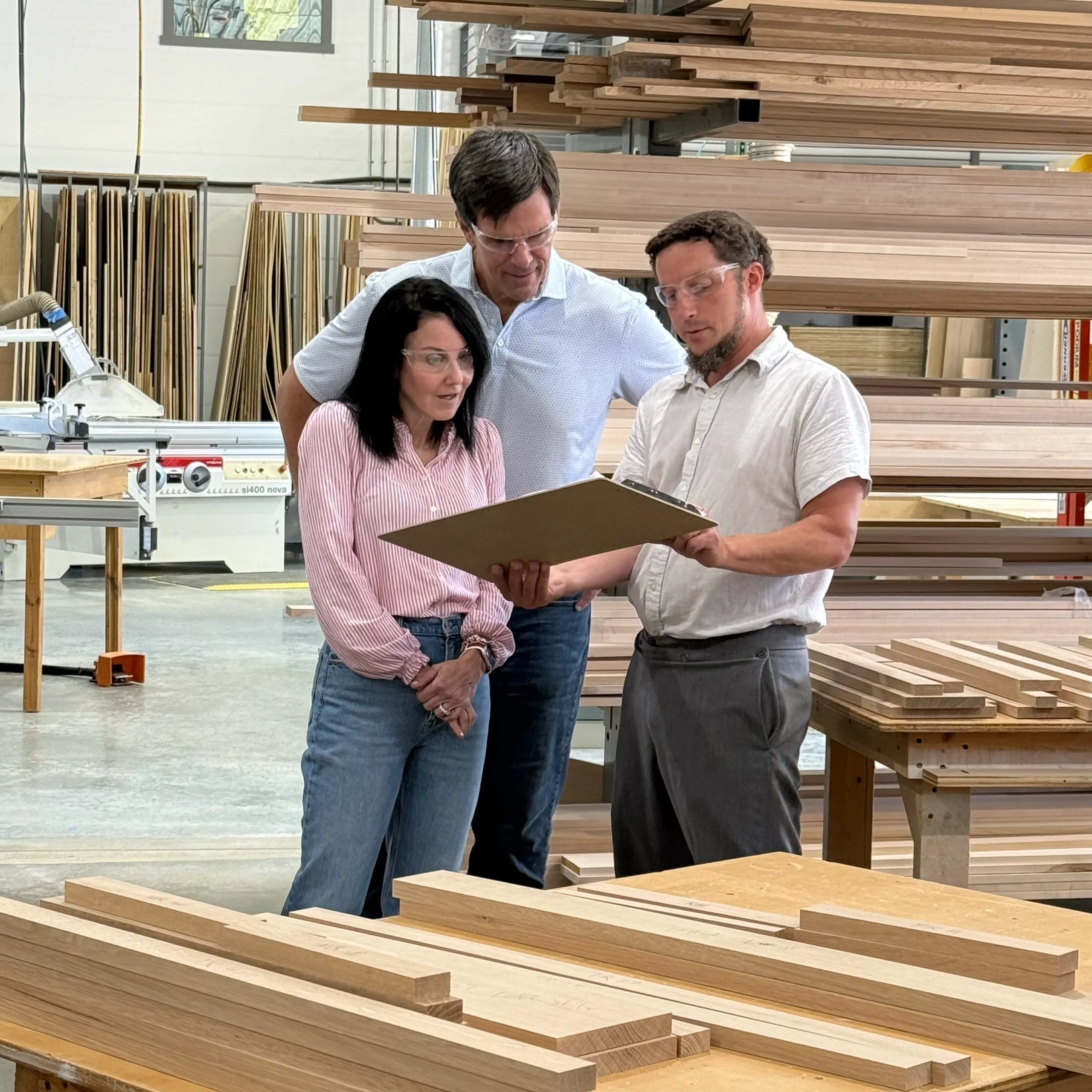 Three people in a woodworking shop examine a large board or sample together. Stacks of wooden planks and woodworking equipment are visible in the background. All individuals are wearing safety glasses.