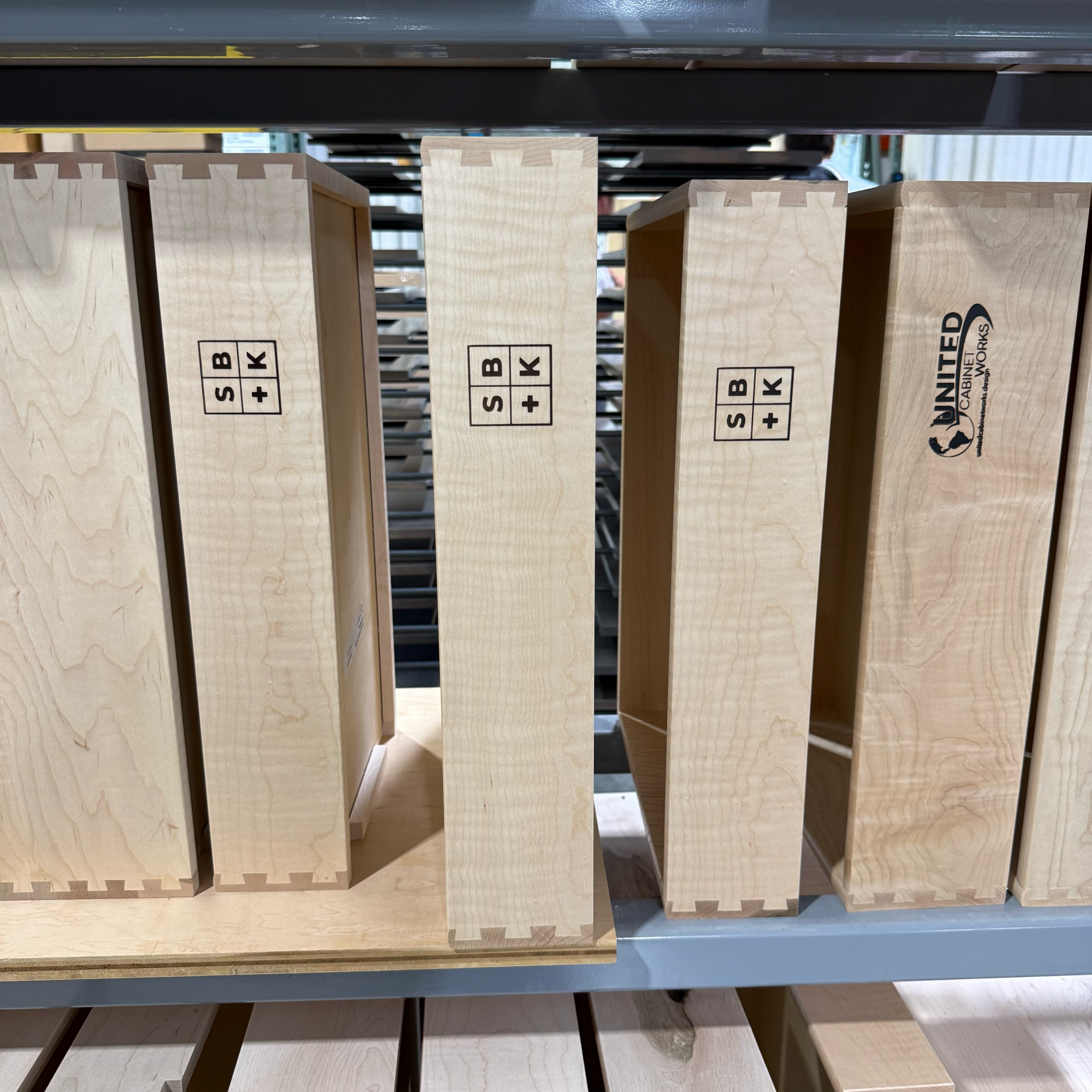 Four wooden drawer sides with dovetail joints are standing vertically on a shelf. Three have SB + K stamped on them, and one has UNITED with an eagle logo. The background shows metal shelves and workshop equipment.